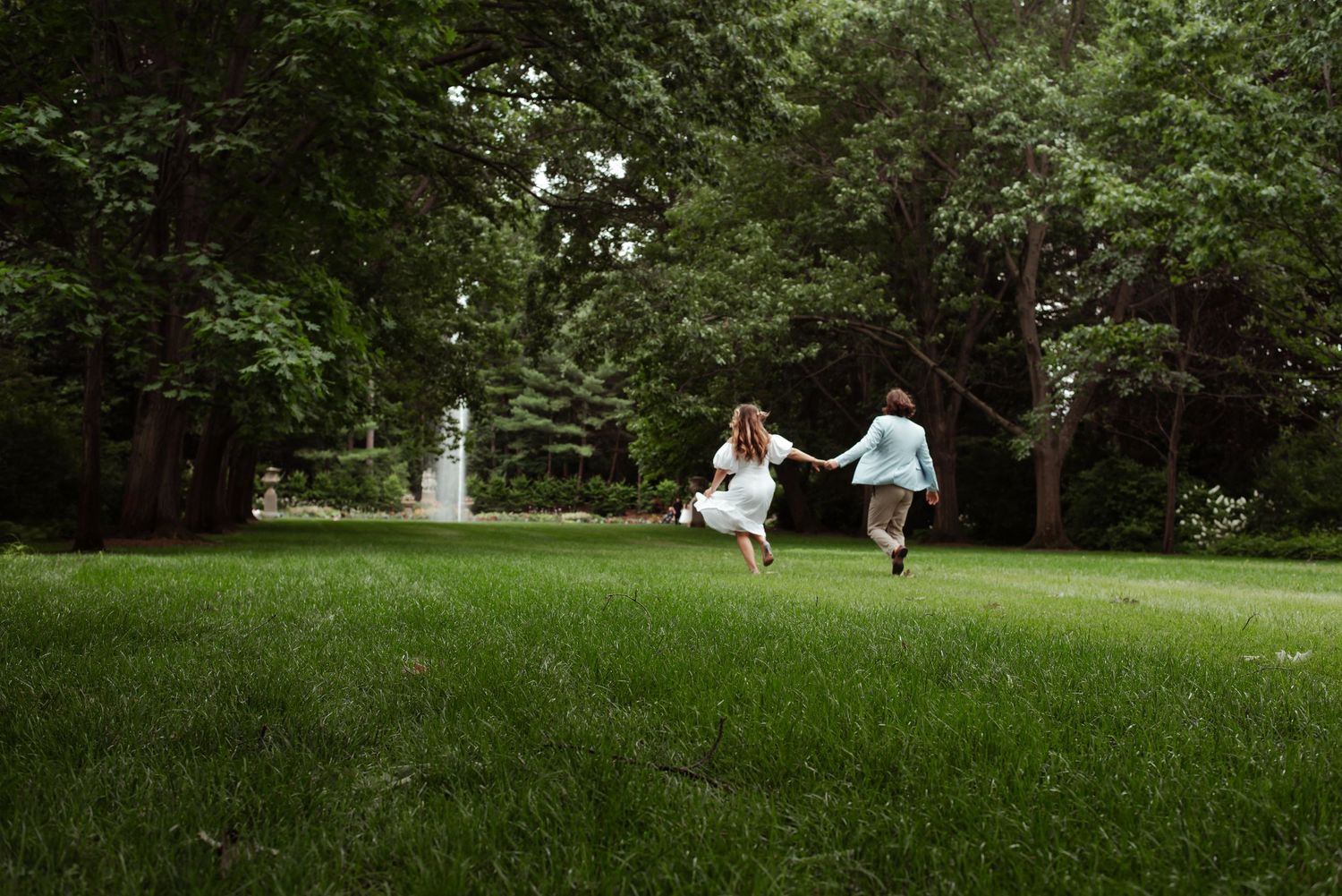 Two children holding hands and running joyfully across a lush green lawn surrounded by tall trees.