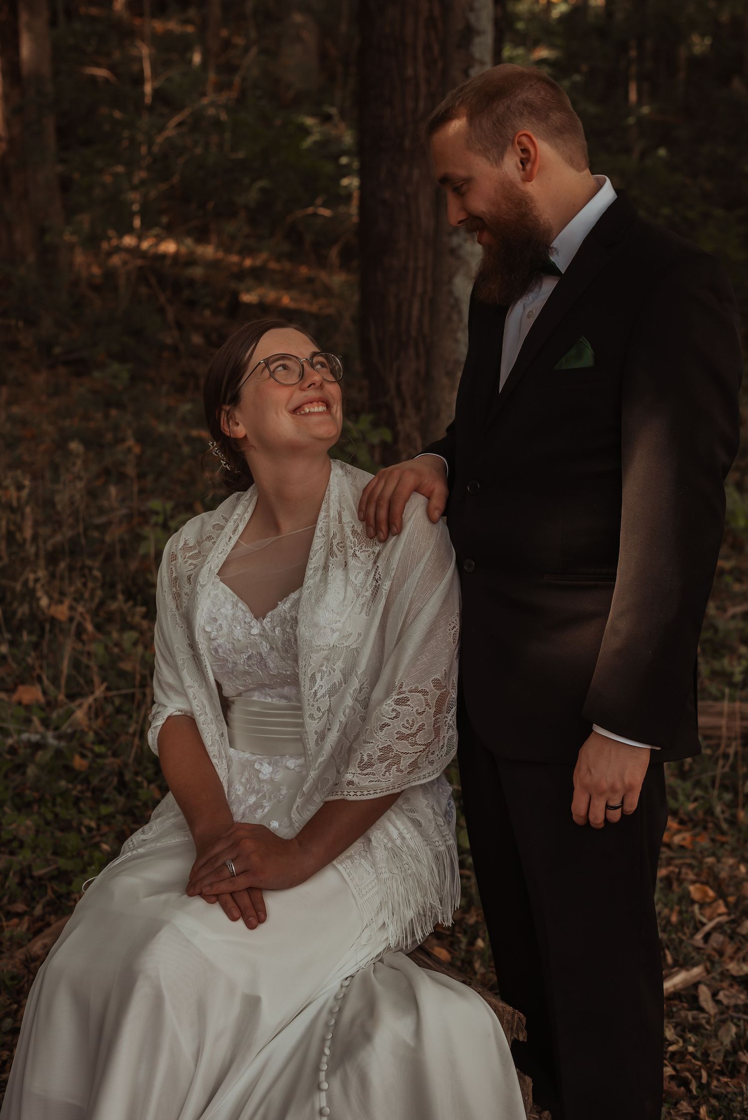 Smiling bride in glasses looks up at bearded groom in forest.
