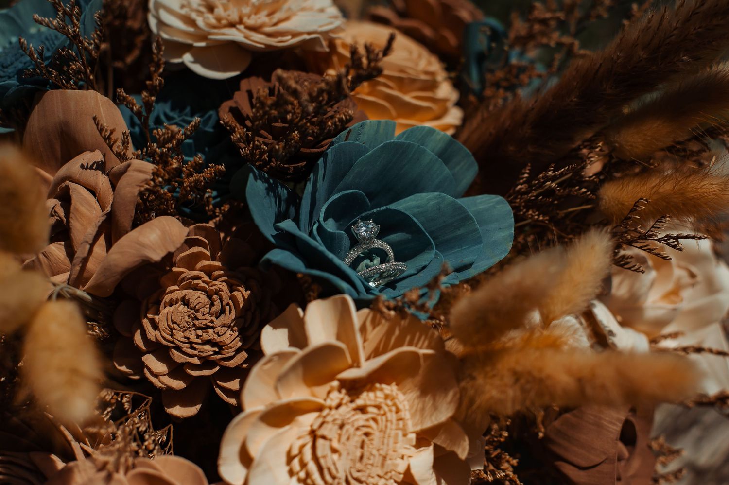 Wedding rings nestled among dried floral arrangement with teal flowers and pine cones.