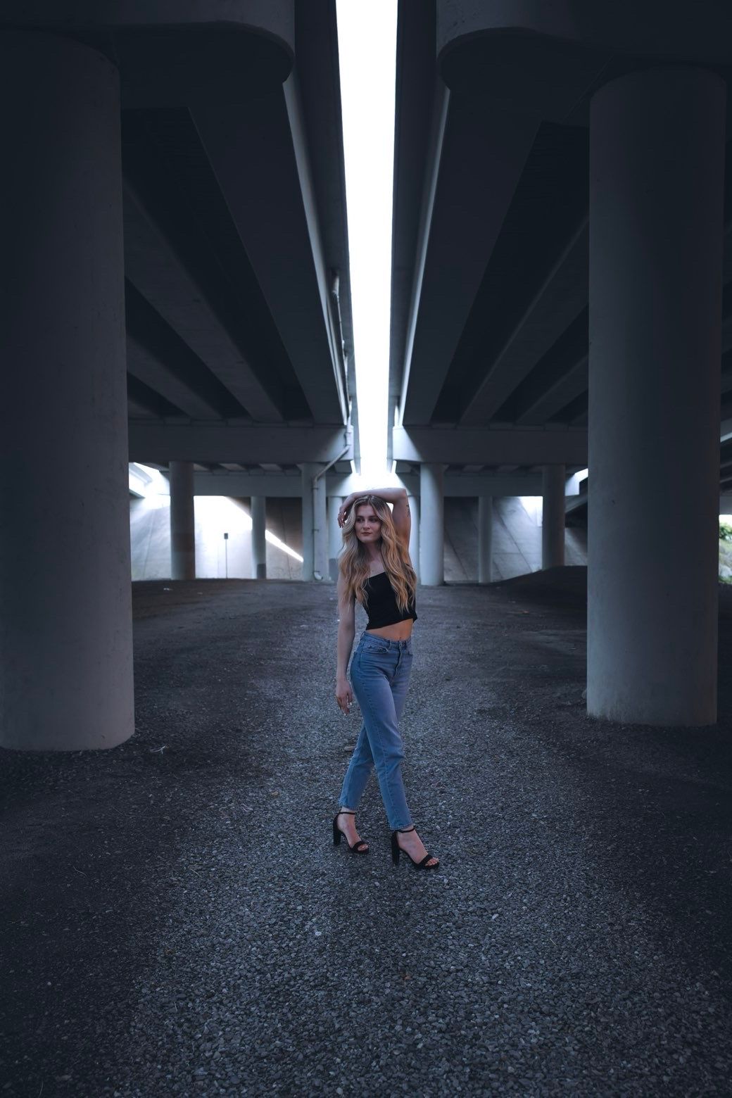 Blonde woman in black crop top and jeans standing under a concrete overpass.