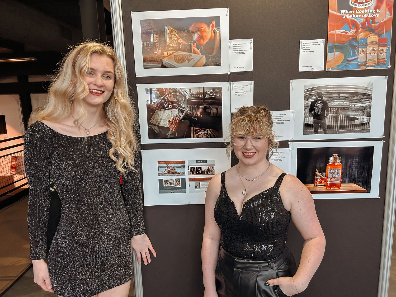 Two women in formal attire posing beside a photography exhibit display.