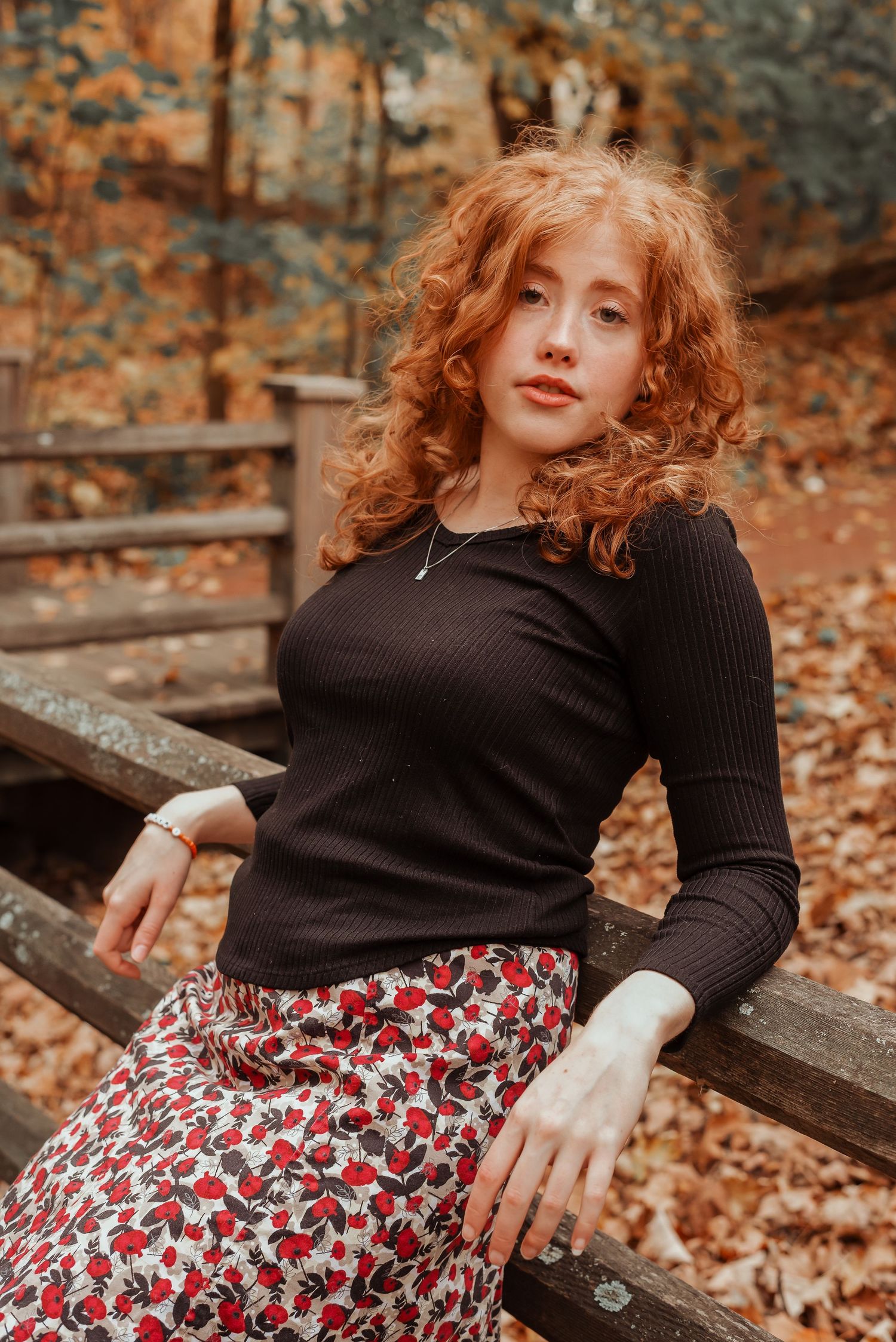 Young woman with curly red hair sits on wooden fence in autumn forest wearing black top and floral skirt.