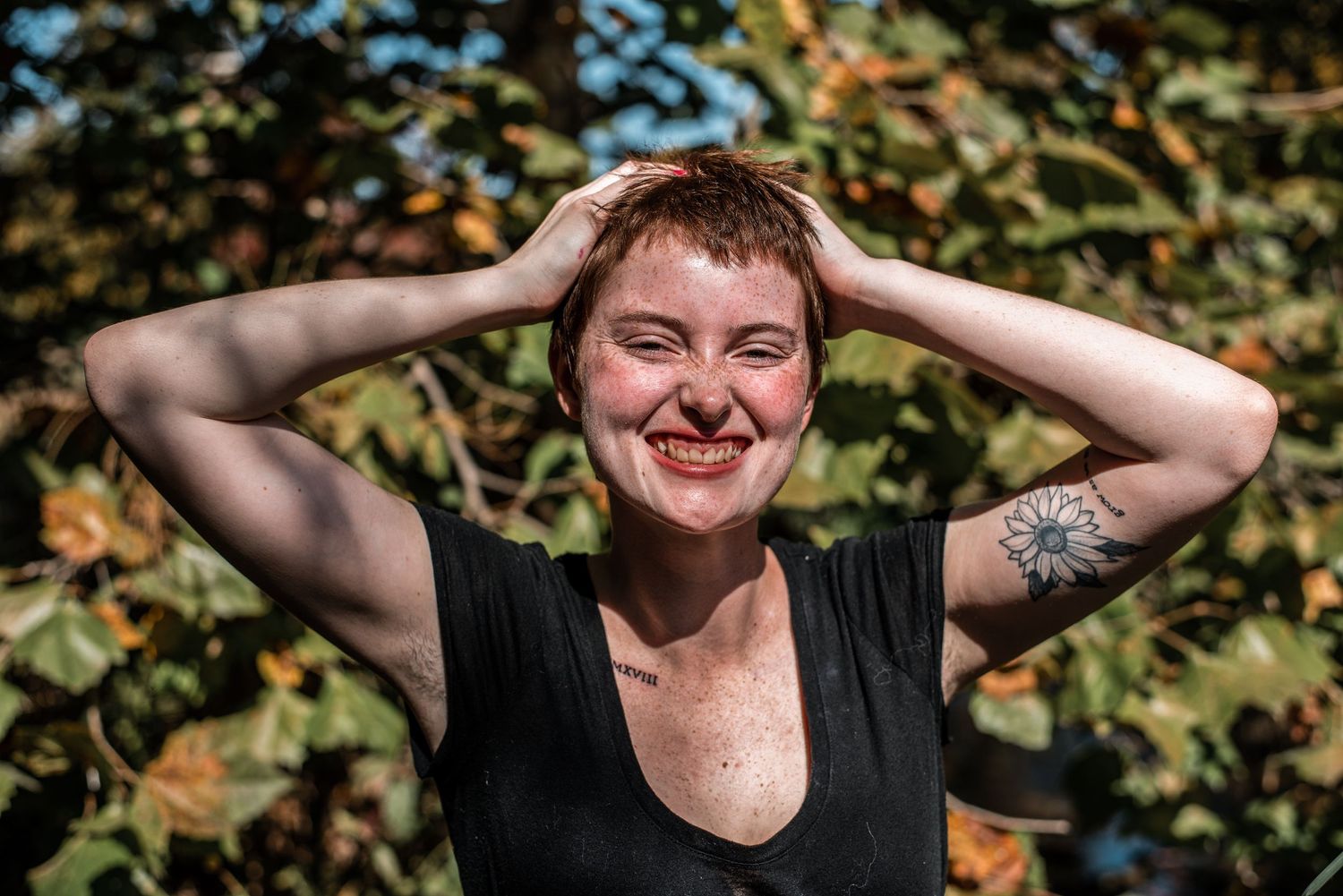 Smiling woman with short hair and tattoo posing outdoors in sunlight.