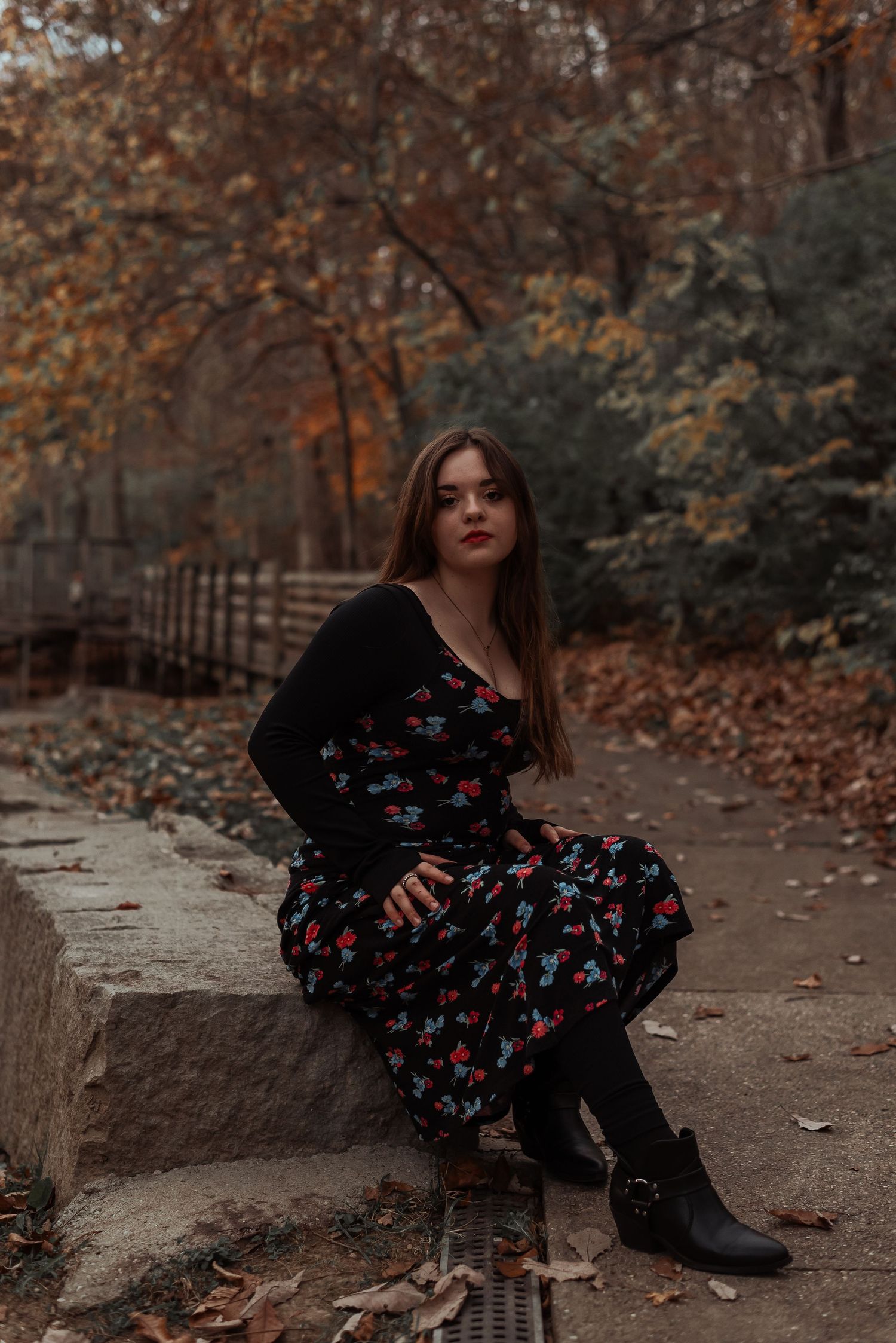 Young woman in floral dress and black boots sits on stone wall in autumn forest park.