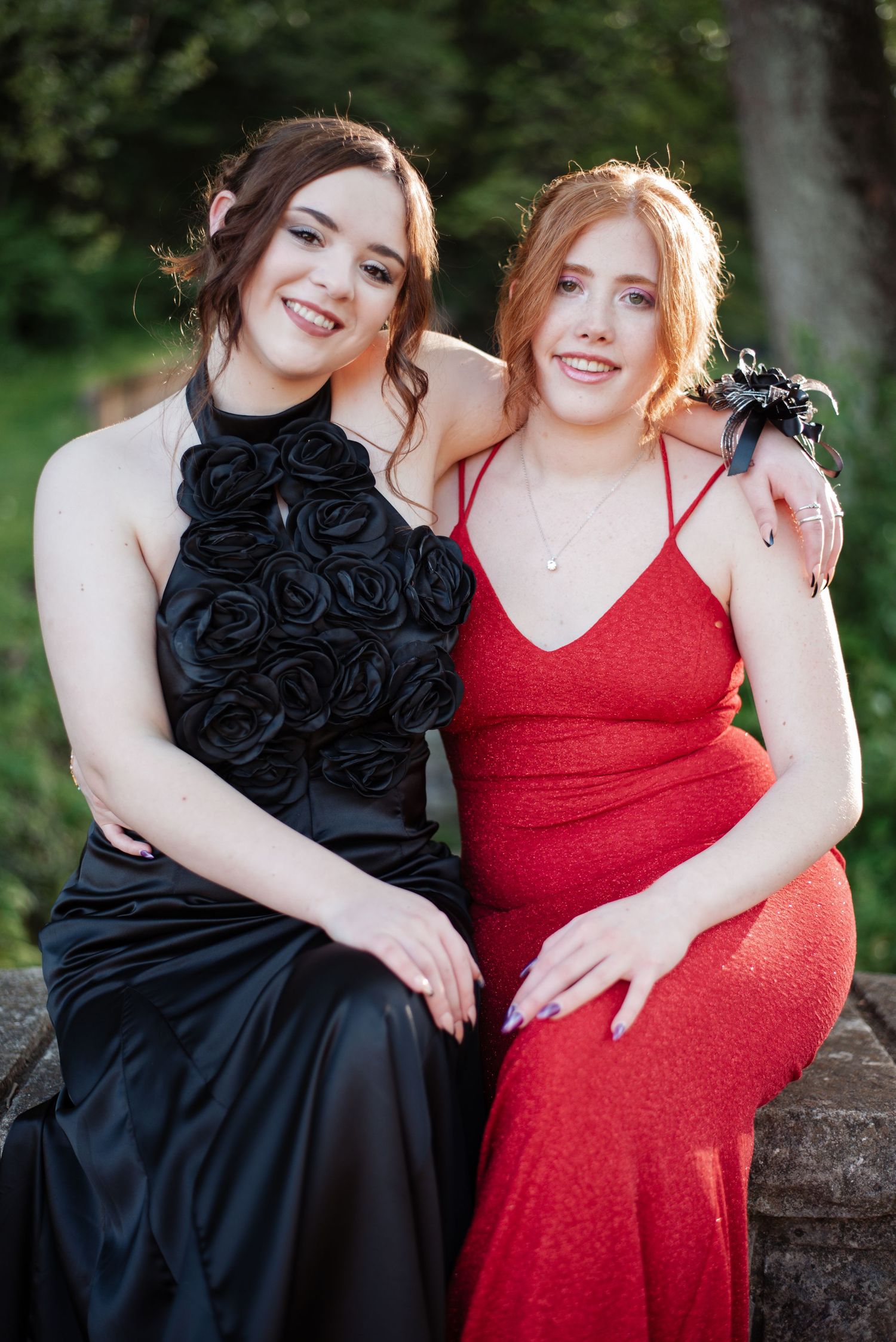 Two smiling prom girls sit together, one in black rose gown and one in red sparkle dress.