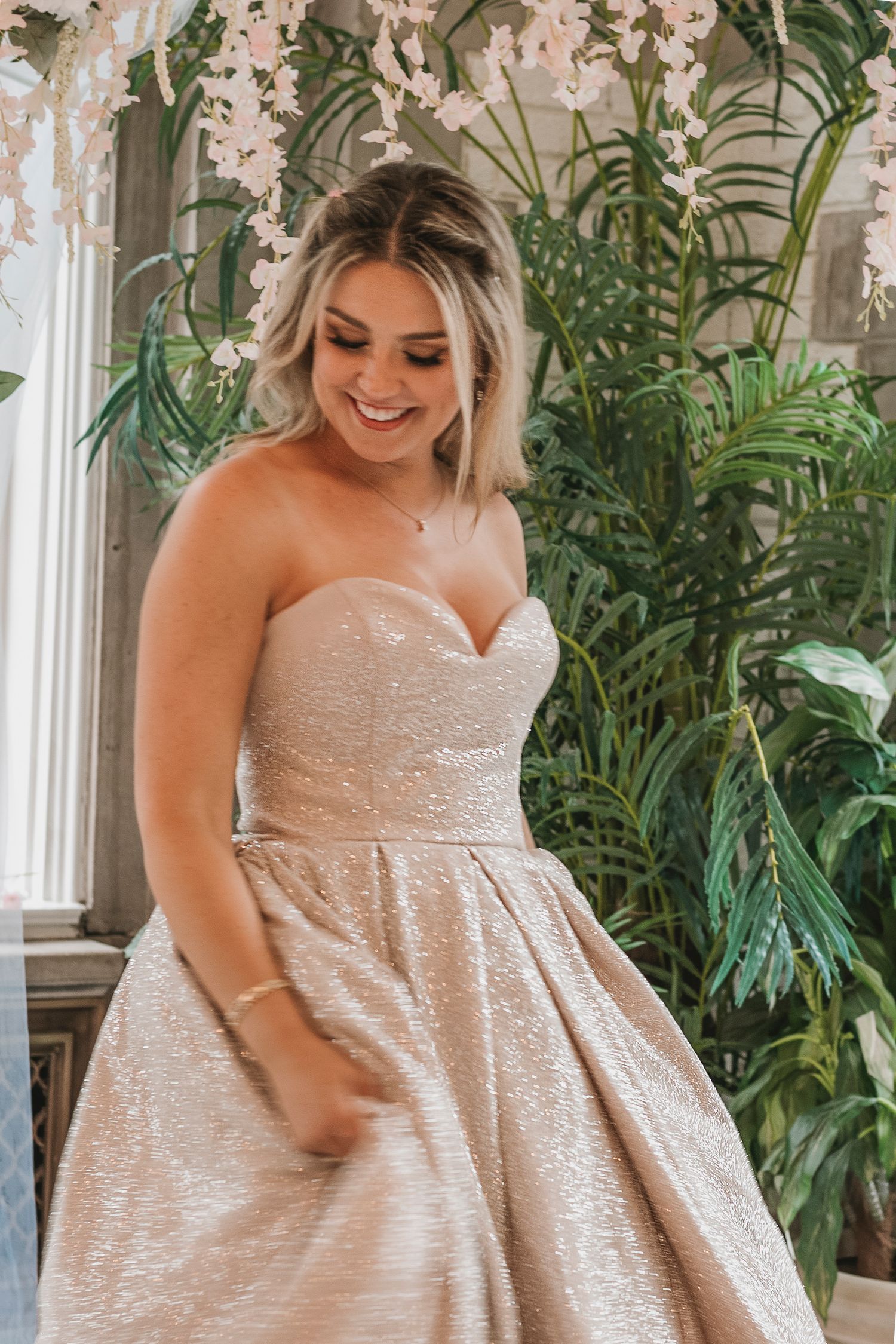Joyful bride in a strapless sparkly champagne ballgown smiling among lush green tropical plants indoors.
