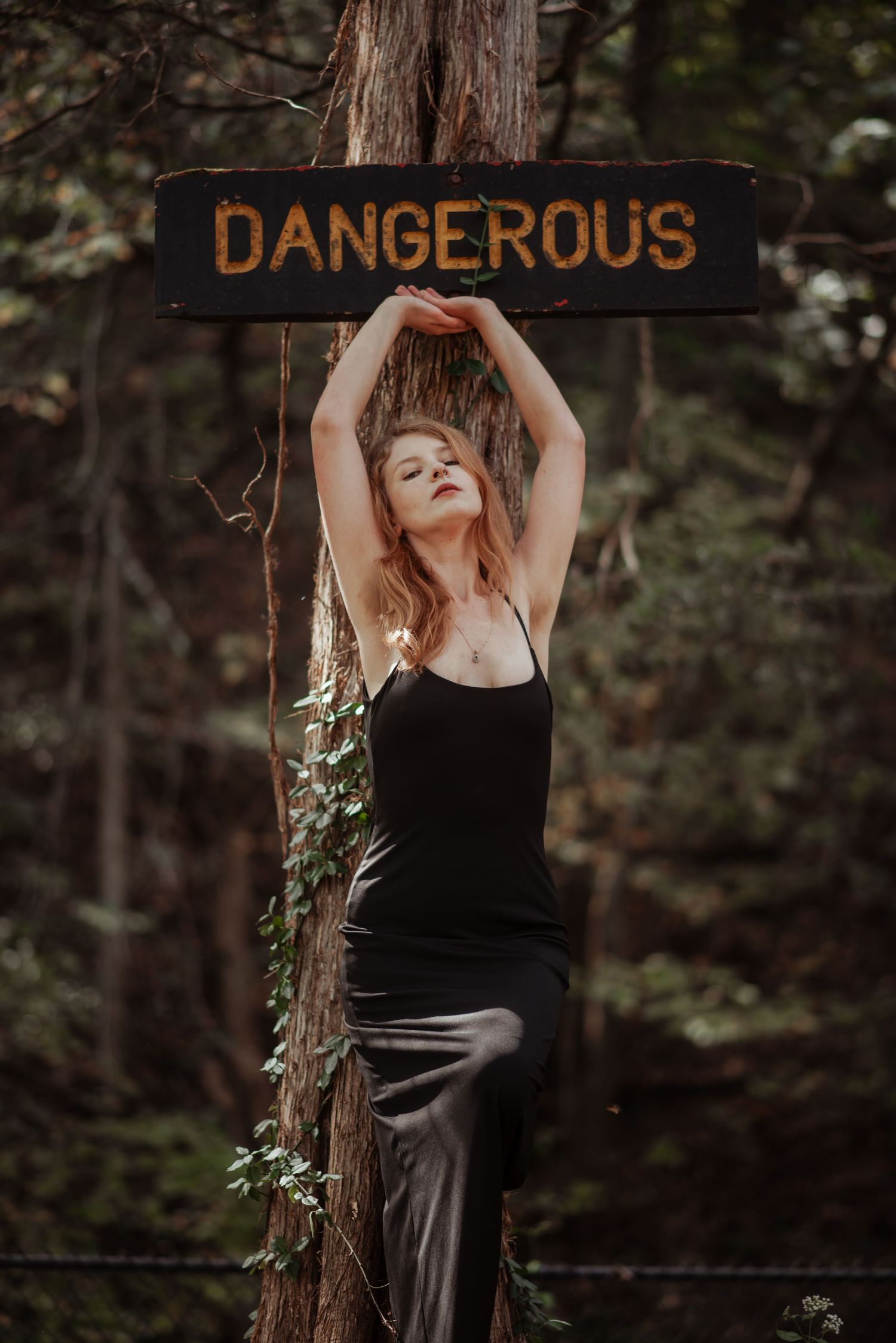 Redhead woman in black outfit raises arms above a Dangerous sign on a forest tree.