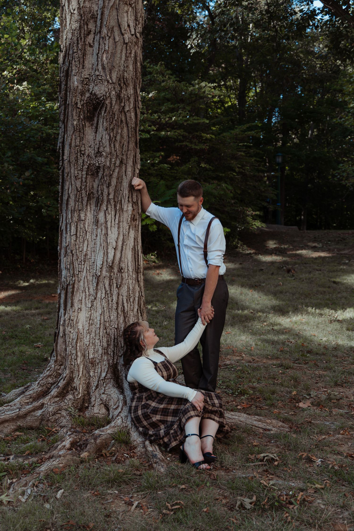 A man leans against a large tree holding a seated woman's hand in a forest park.