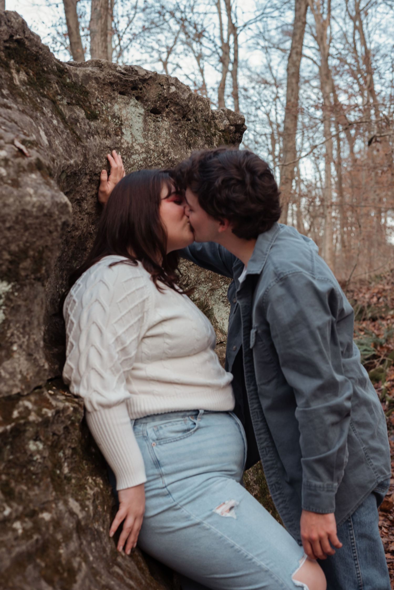 A couple shares a tender kiss against a mossy boulder surrounded by bare autumn trees.