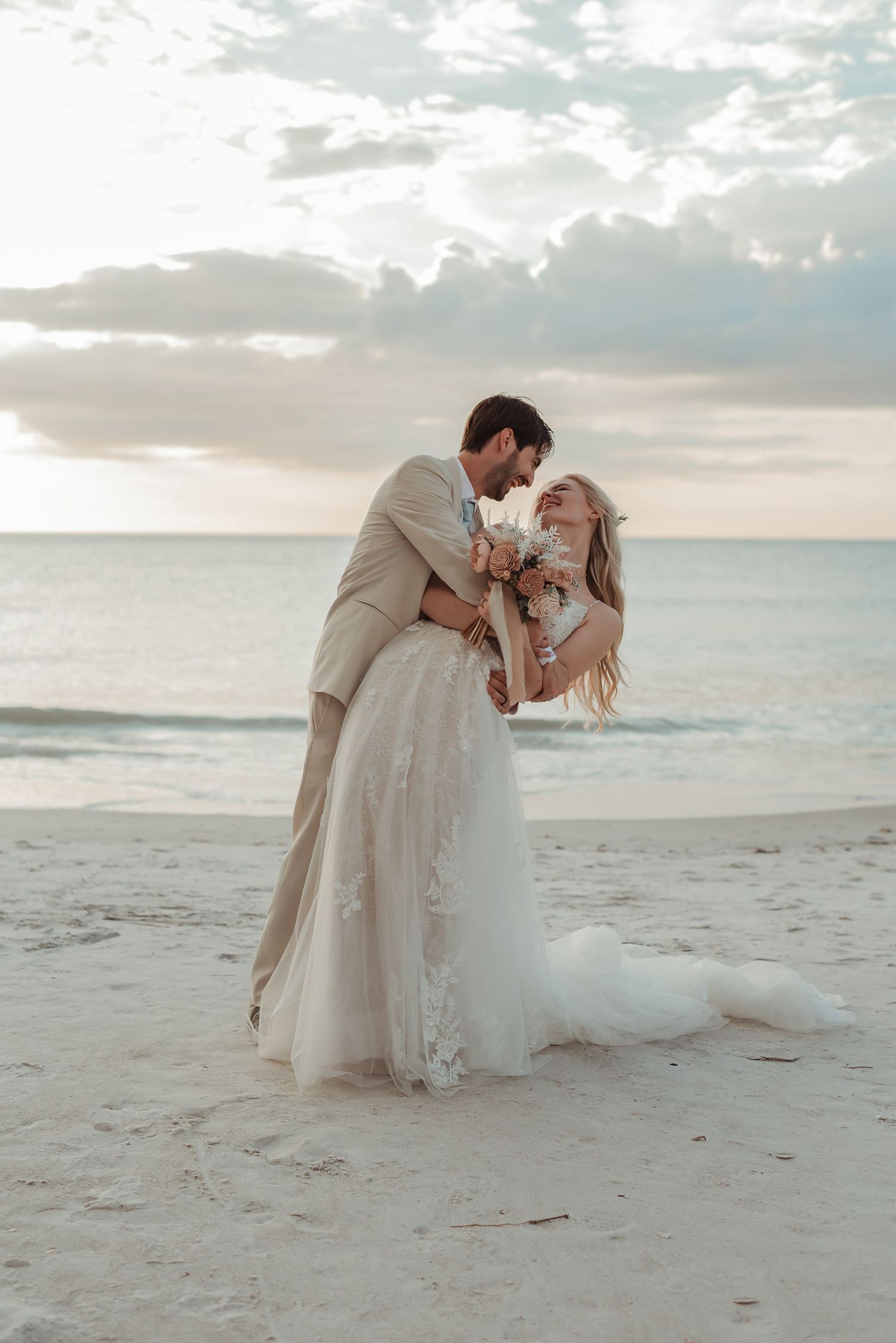 A groom dips his bride for a kiss on a sandy beach during a romantic sunset wedding.
