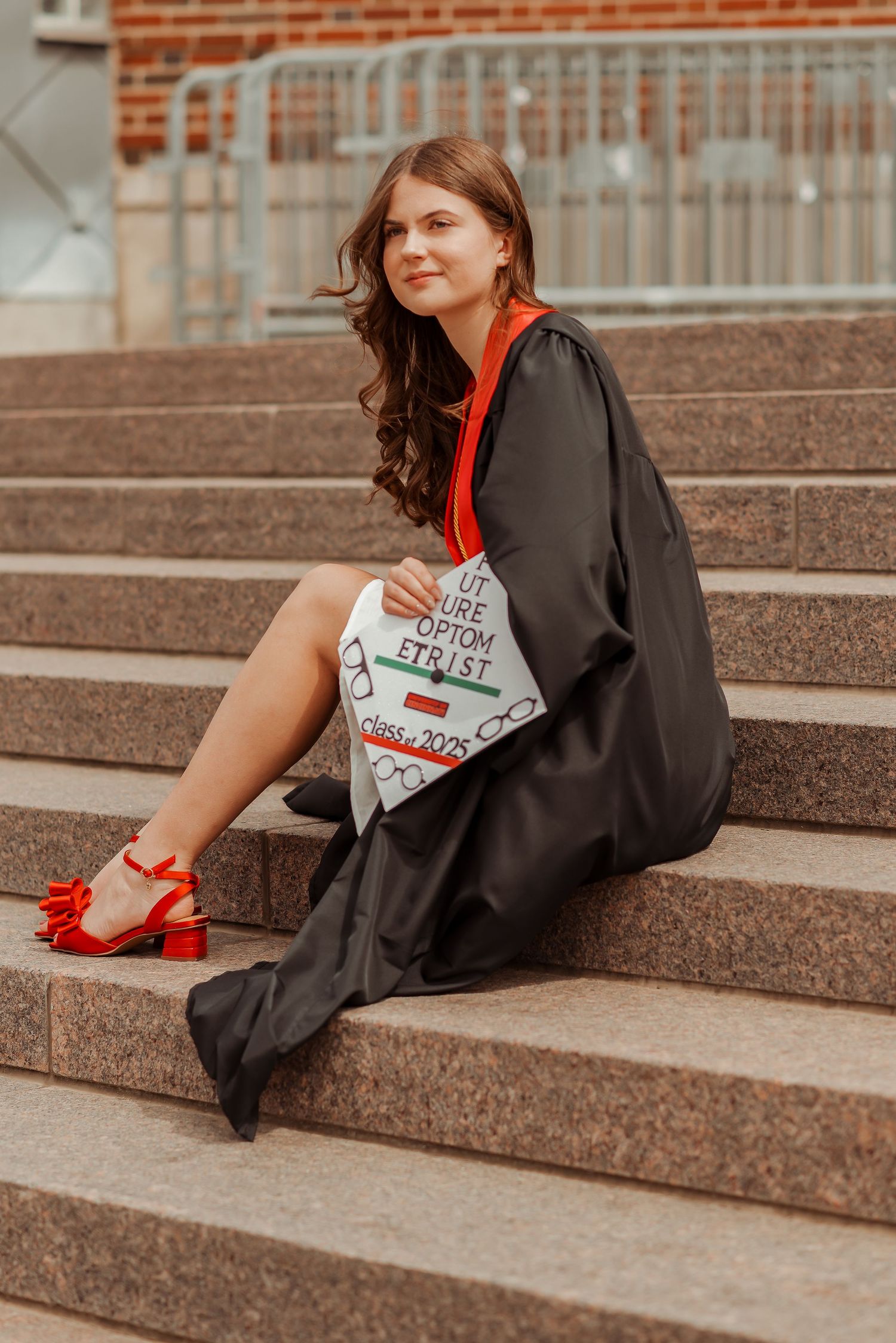 Graduate in black gown sitting on stone steps holding a decorated optometry graduation cap, Class of 2025.