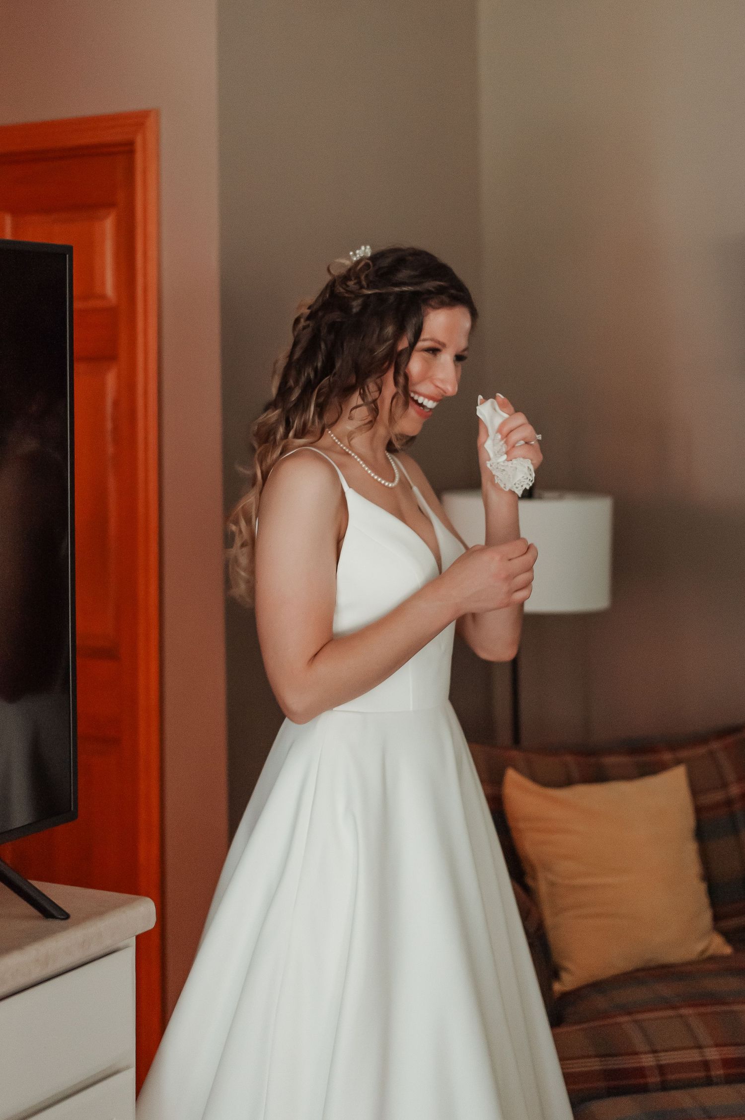 Smiling bride in elegant white V-neck wedding gown holding bouquet indoors before ceremony.