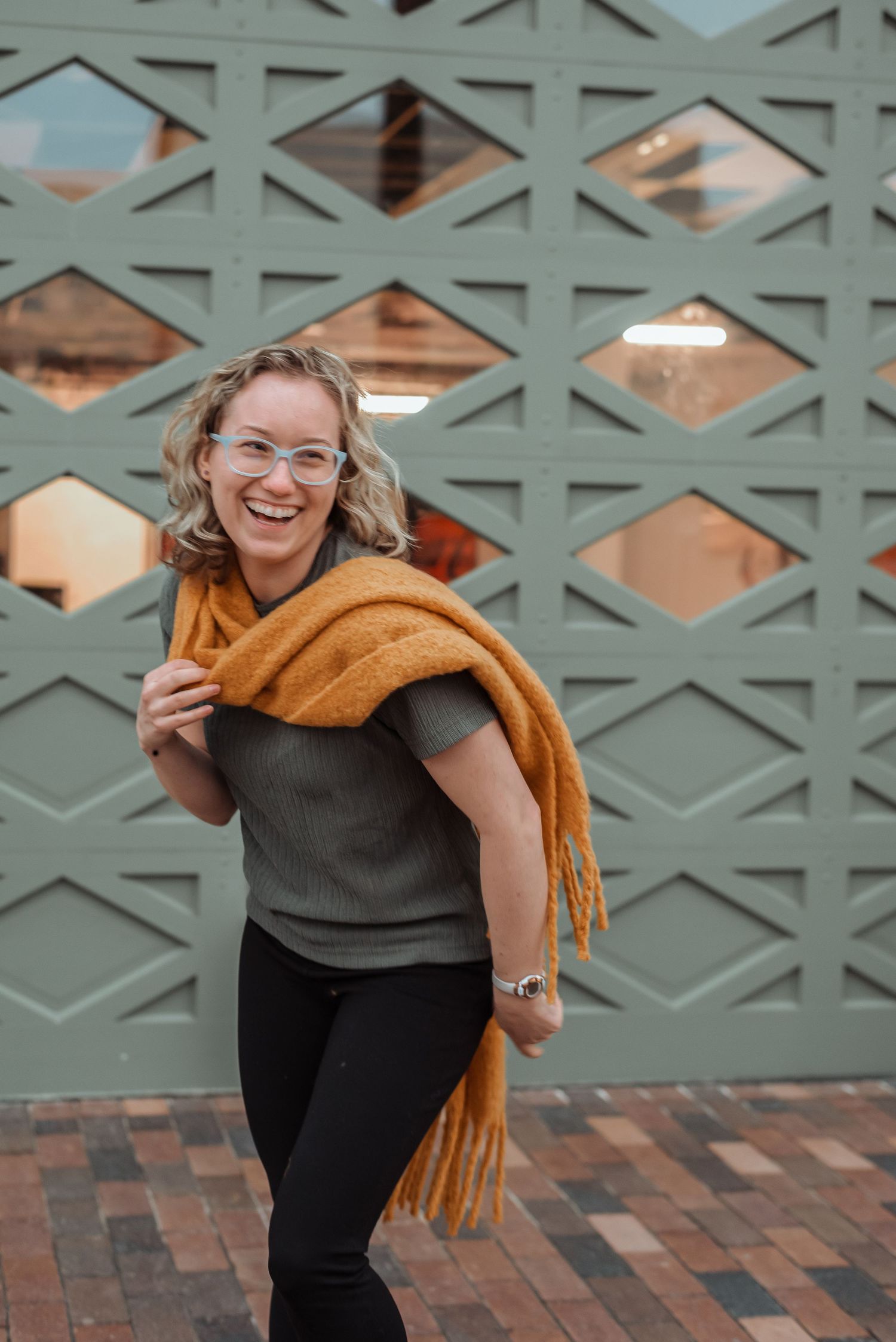 Laughing young woman with glasses wearing a mustard scarf posed in front of a decorative green gate.