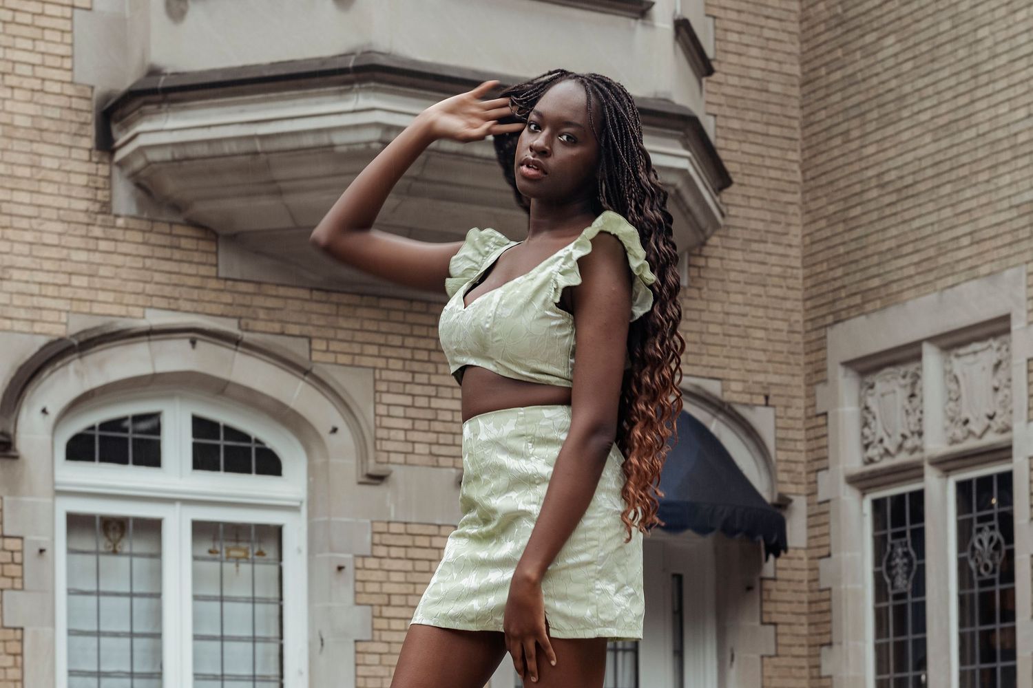 Young woman in light green crop top and mini skirt posing outside brick building.