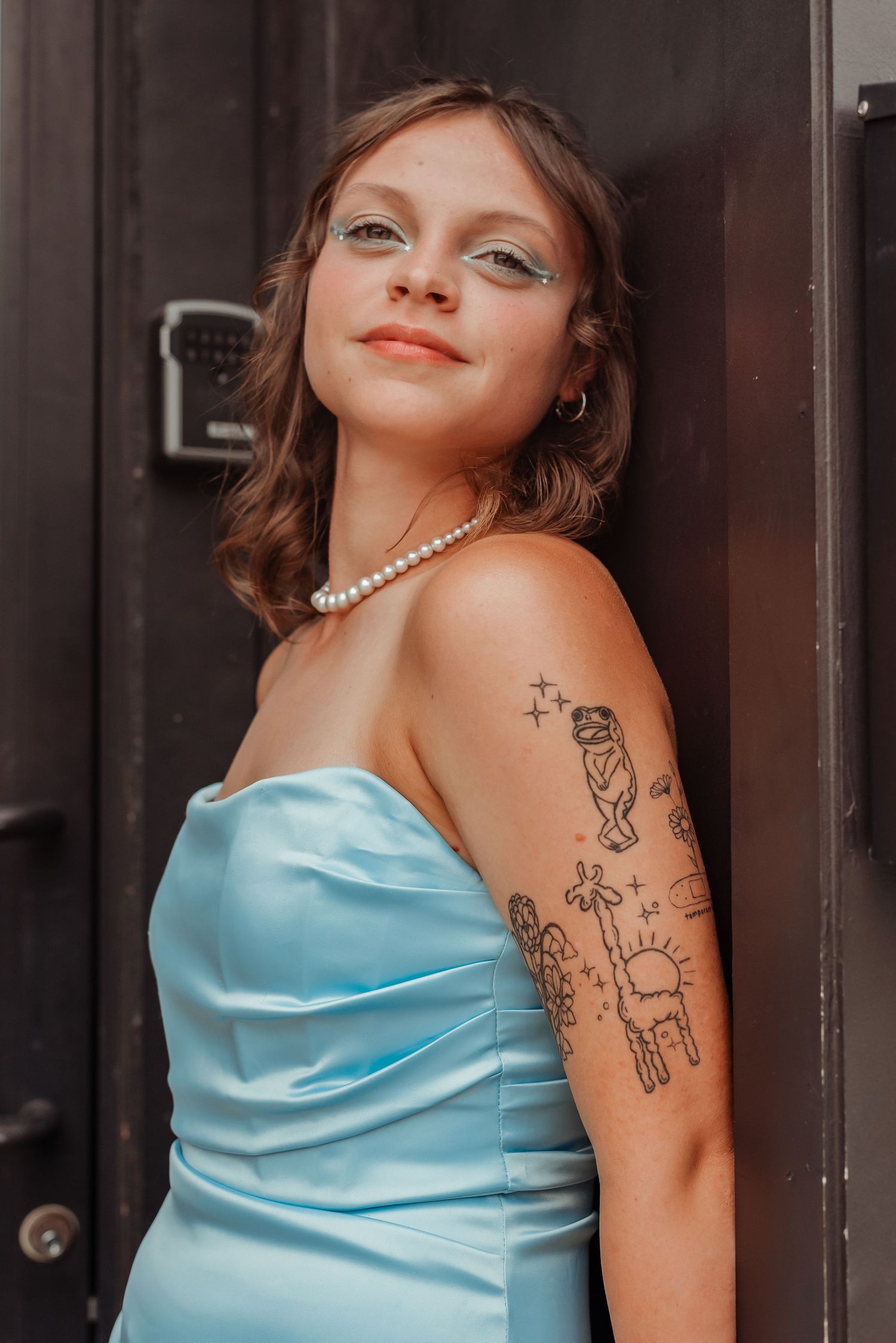 Woman in blue strapless dress with arm tattoos and pearl necklace leaning against dark door.