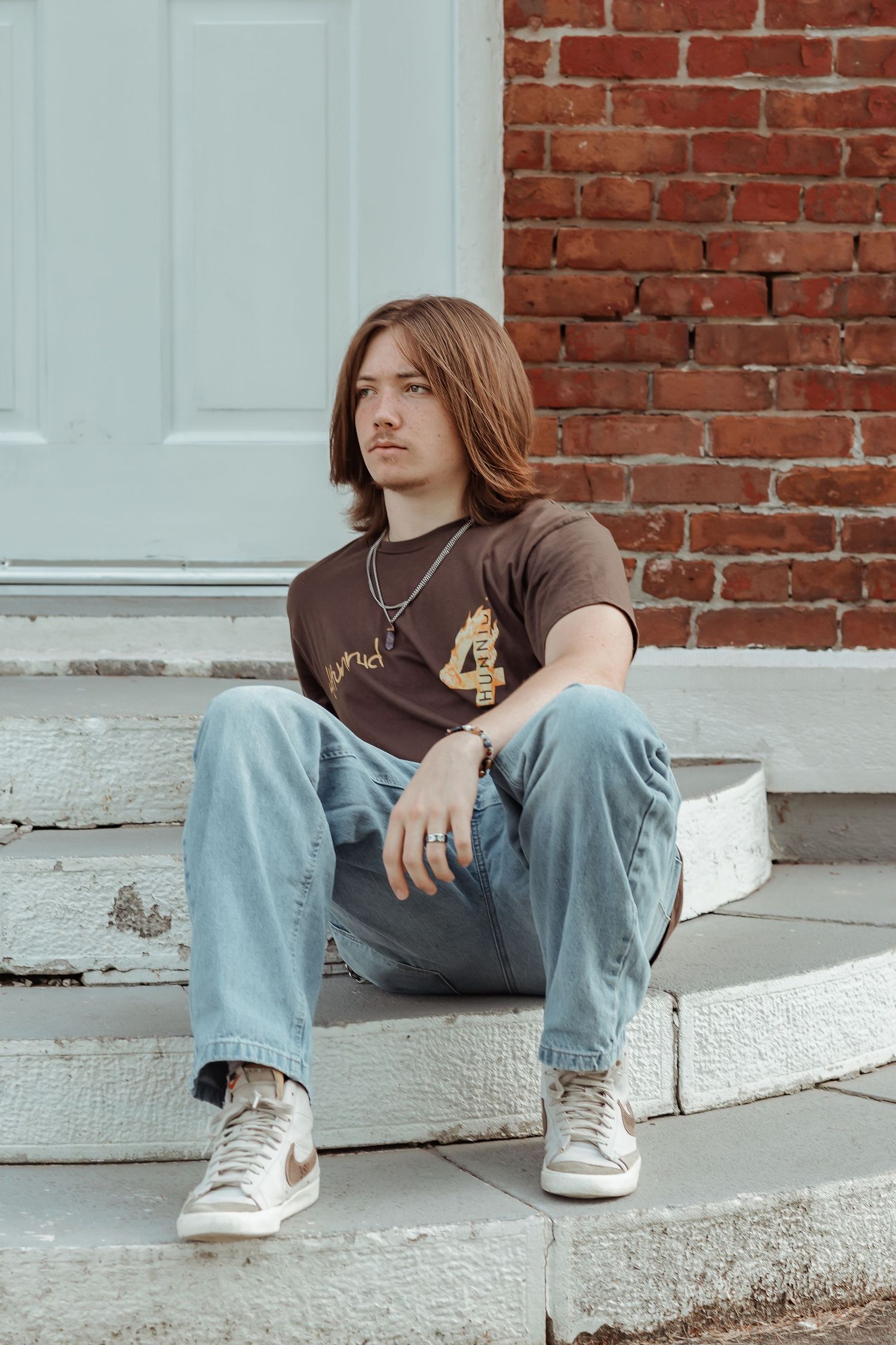 Young man with long hair sitting on stone steps in jeans and brown tee shirt against brick wall.