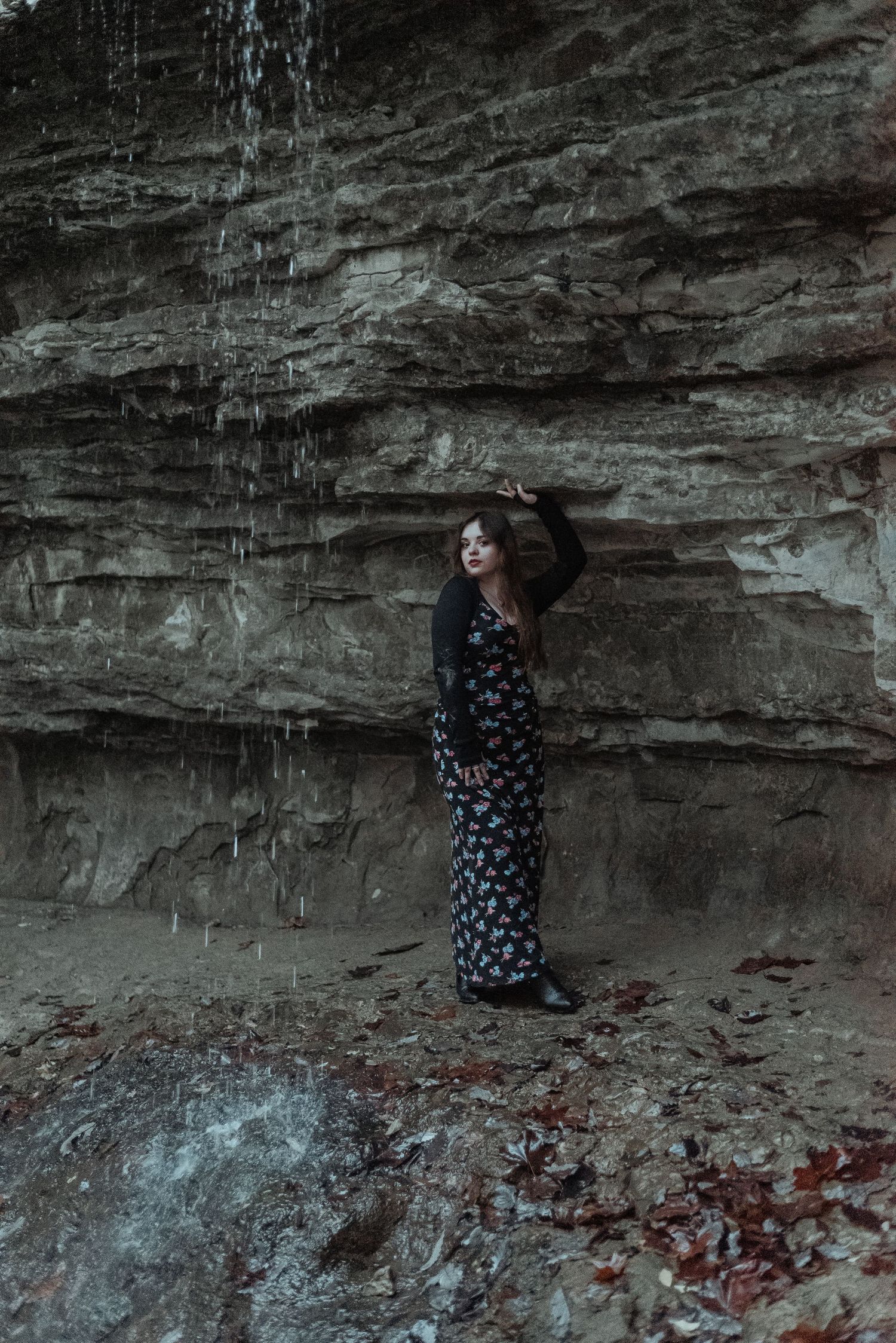 Woman in black floral maxi dress standing near rocky waterfall cliff wall outdoors.