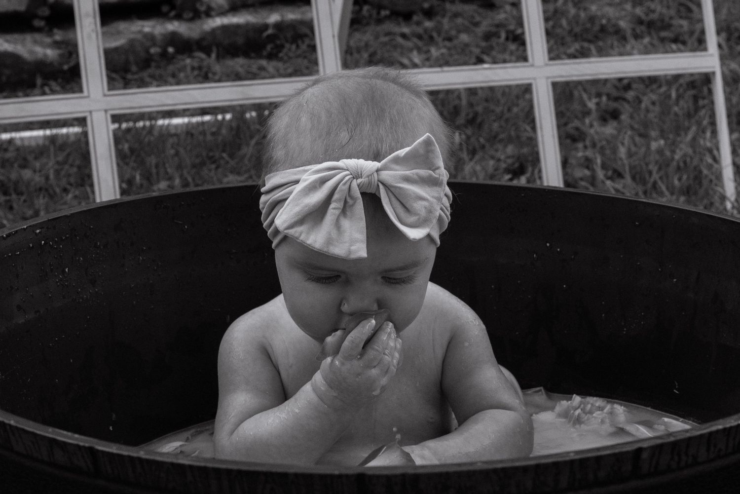 Black and white photo of baby with bow headband splashing in outdoor tub during photo session.