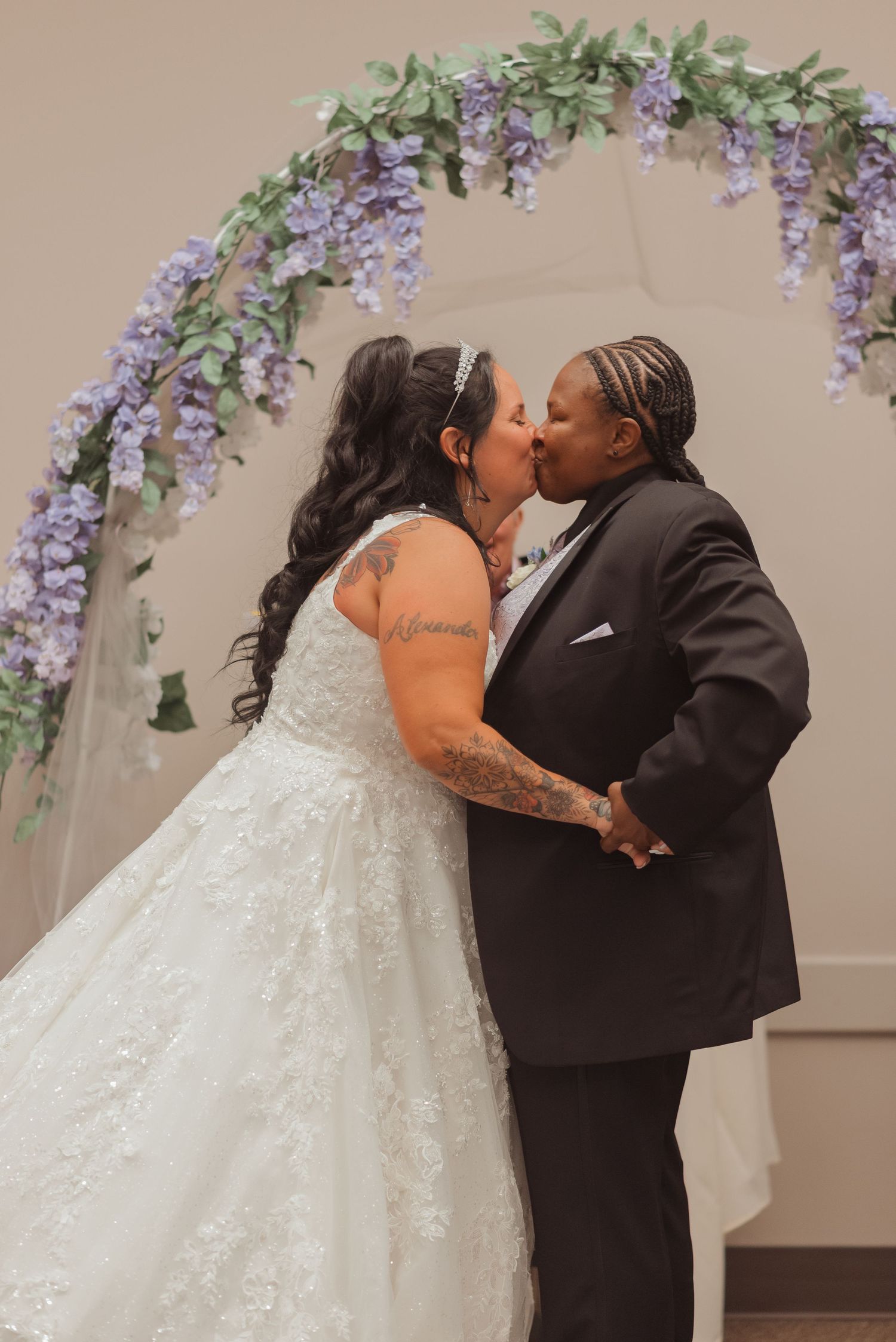 Two brides share first kiss at altar adorned with purple wisteria floral arch, one in white gown and one in black suit.