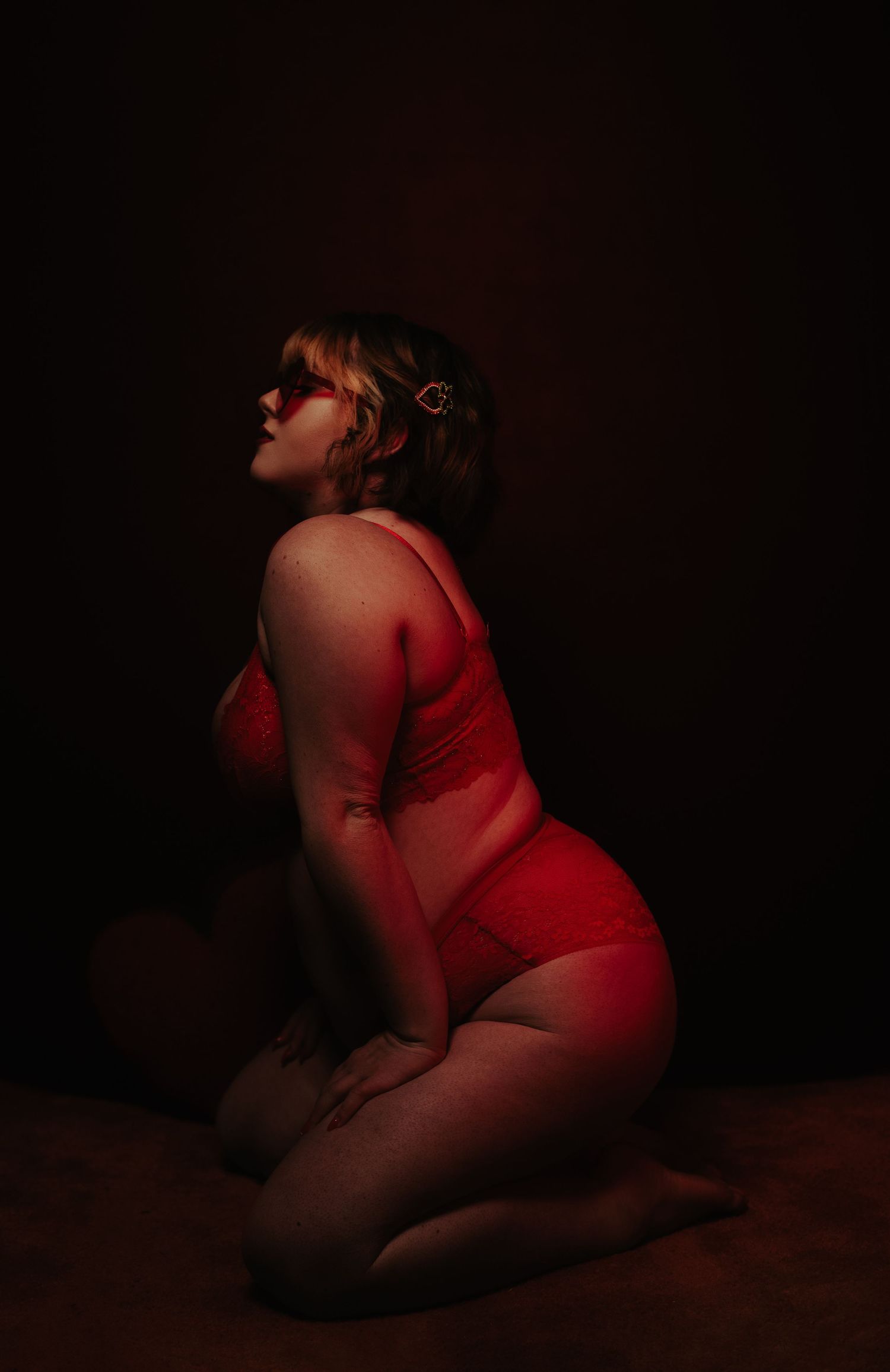 Plus-size woman in red bodysuit kneeling in dramatic dark studio lighting.