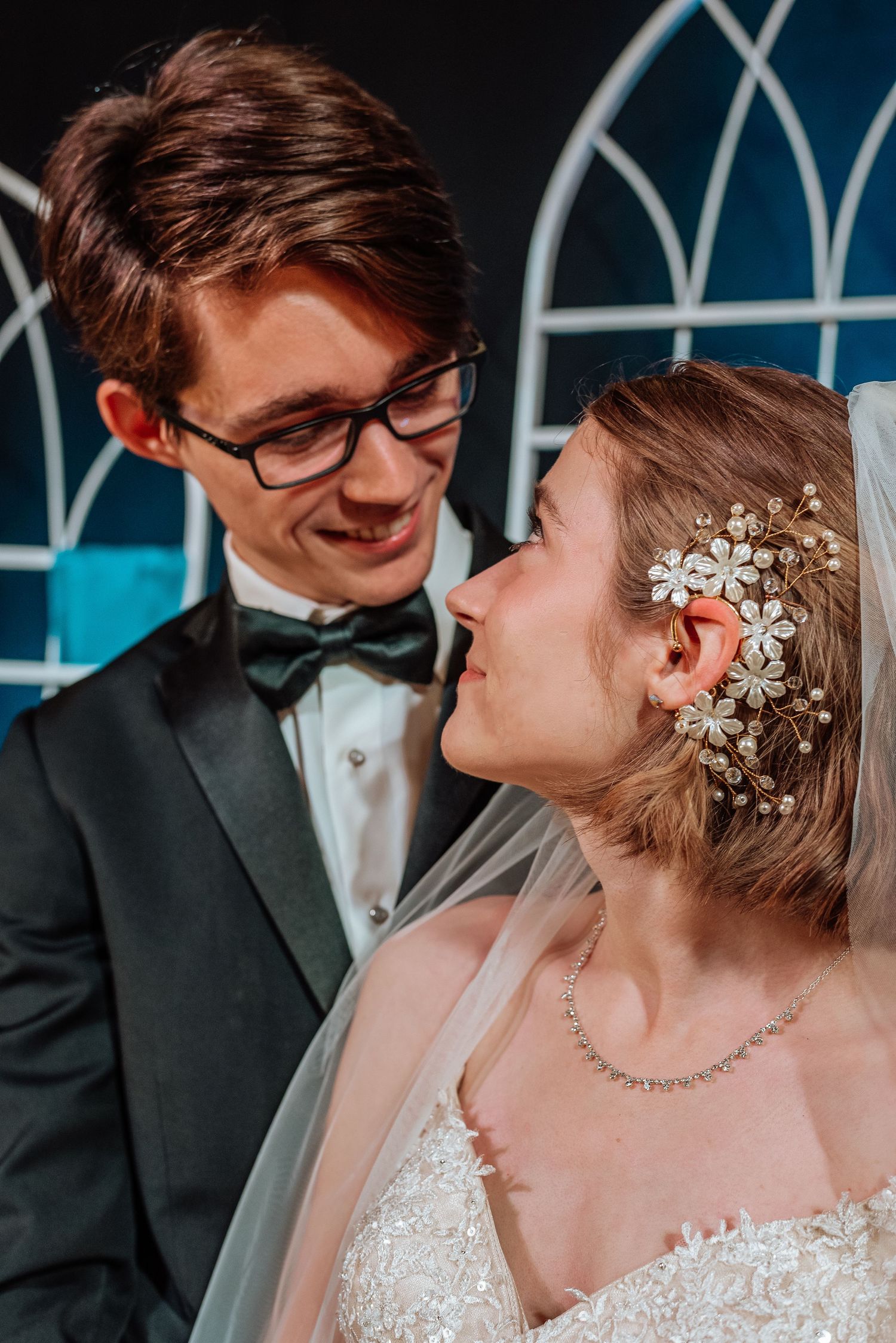 A smiling groom in a tuxedo gazes at his bride wearing a floral hair piece and veil on their wedding day.