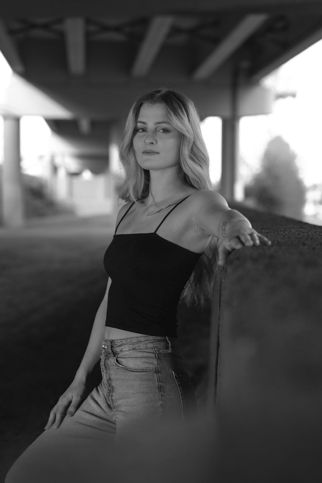 Black and white portrait of a blonde woman in a black top leaning against a wall under a bridge.