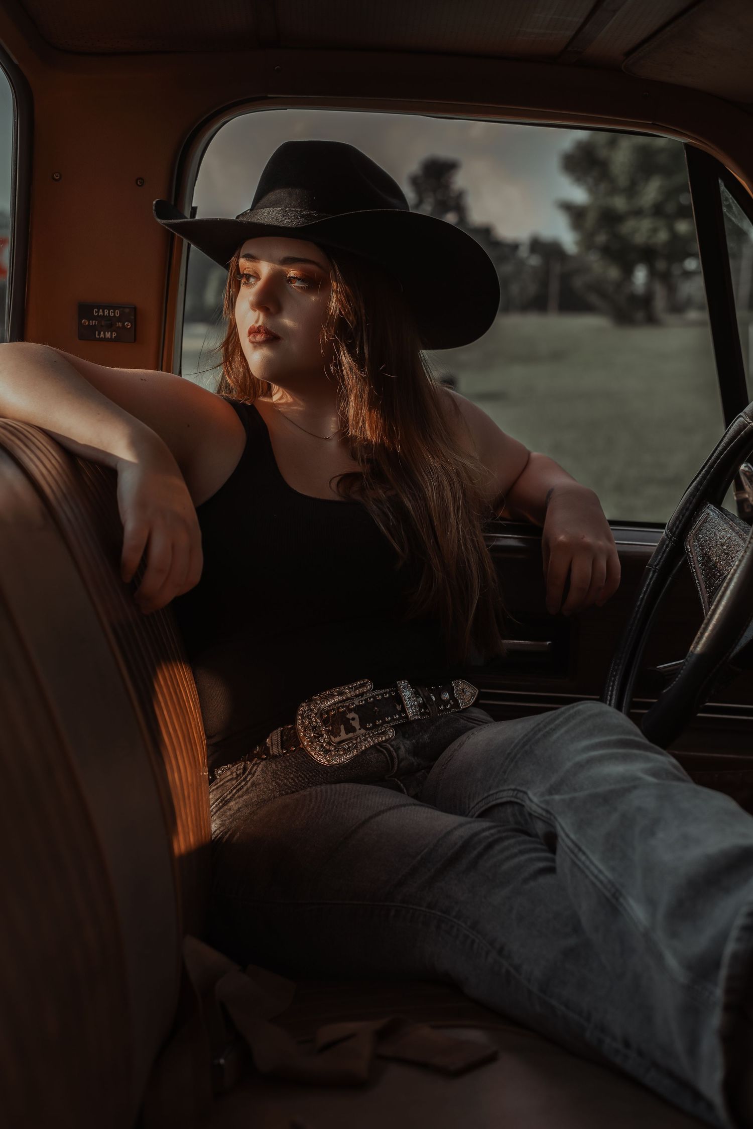 Young woman in black tank top, cowboy hat, and jeans poses inside vintage truck cab at golden hour.