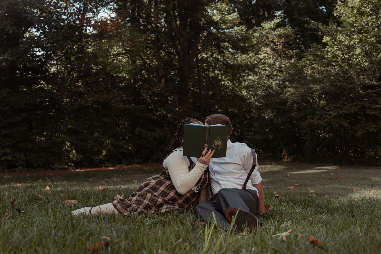 A couple sits on grass in a park hiding their faces behind a green book together.