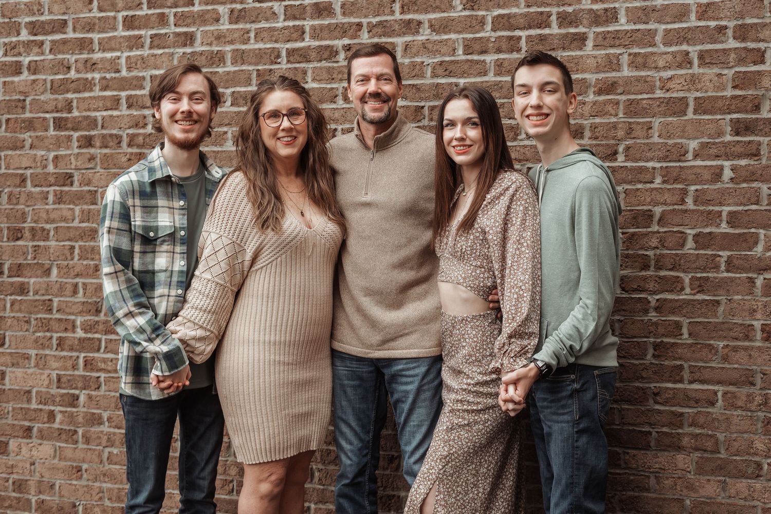 Happy family of five poses together smiling in front of a rustic brick wall for a family portrait.