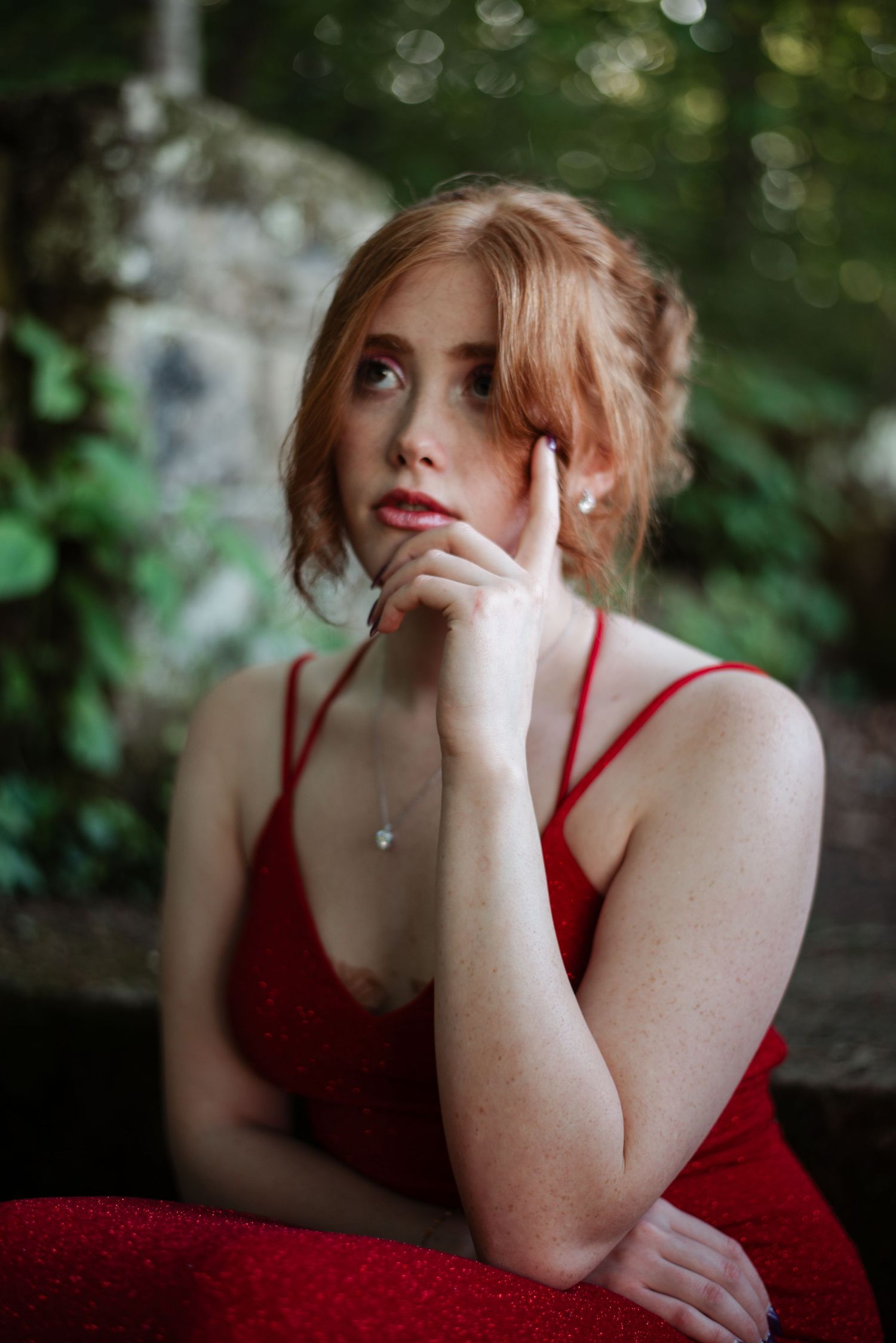 Young woman in red prom dress poses thoughtfully with hand on chin in outdoor setting.