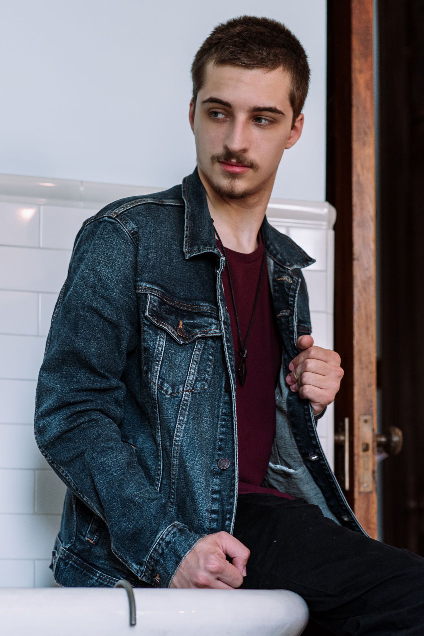 Young man in denim jacket and burgundy shirt sitting casually indoors.