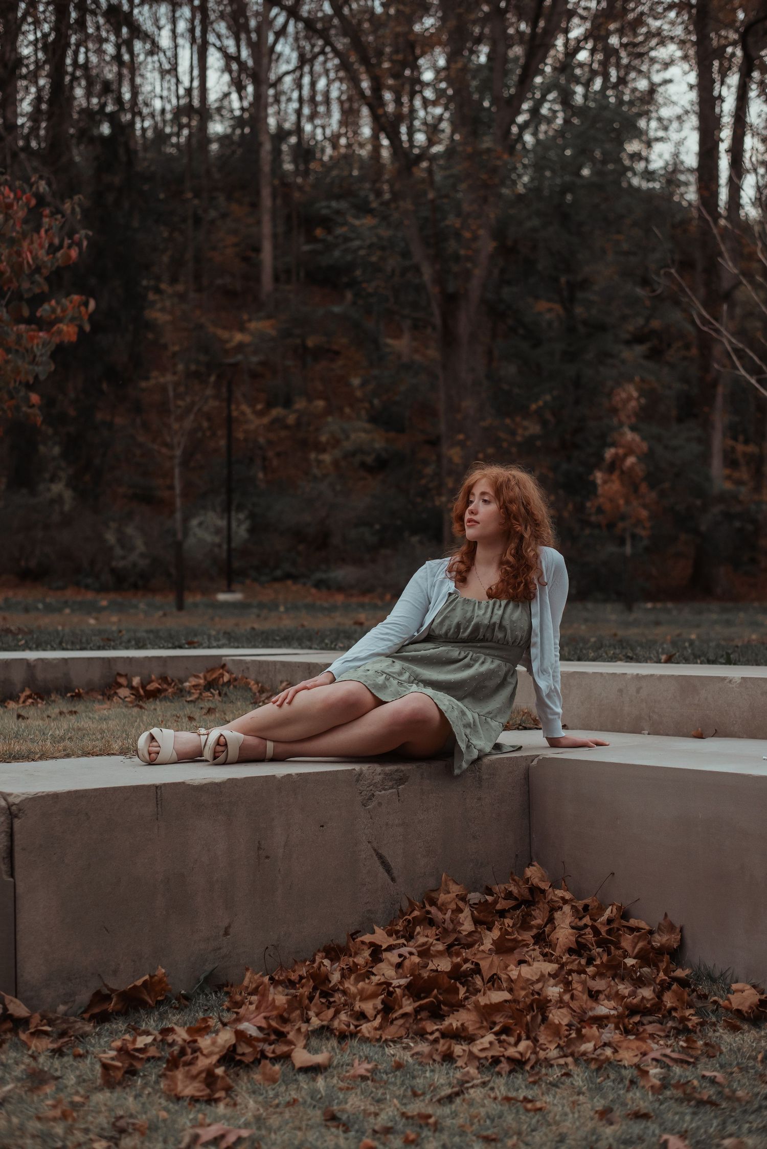 Young woman with curly red hair in green dress sitting on concrete ledge with autumn leaves.