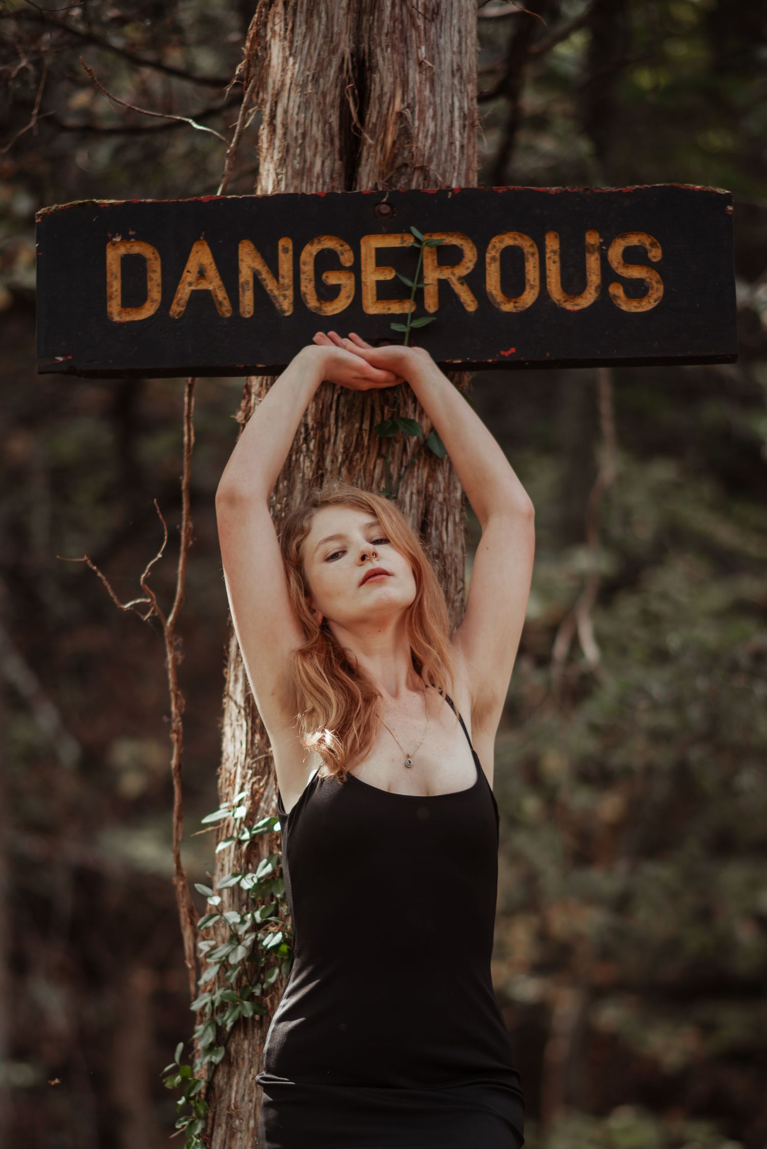 Woman in black dress poses under a rustic Dangerous sign attached to a forest tree.