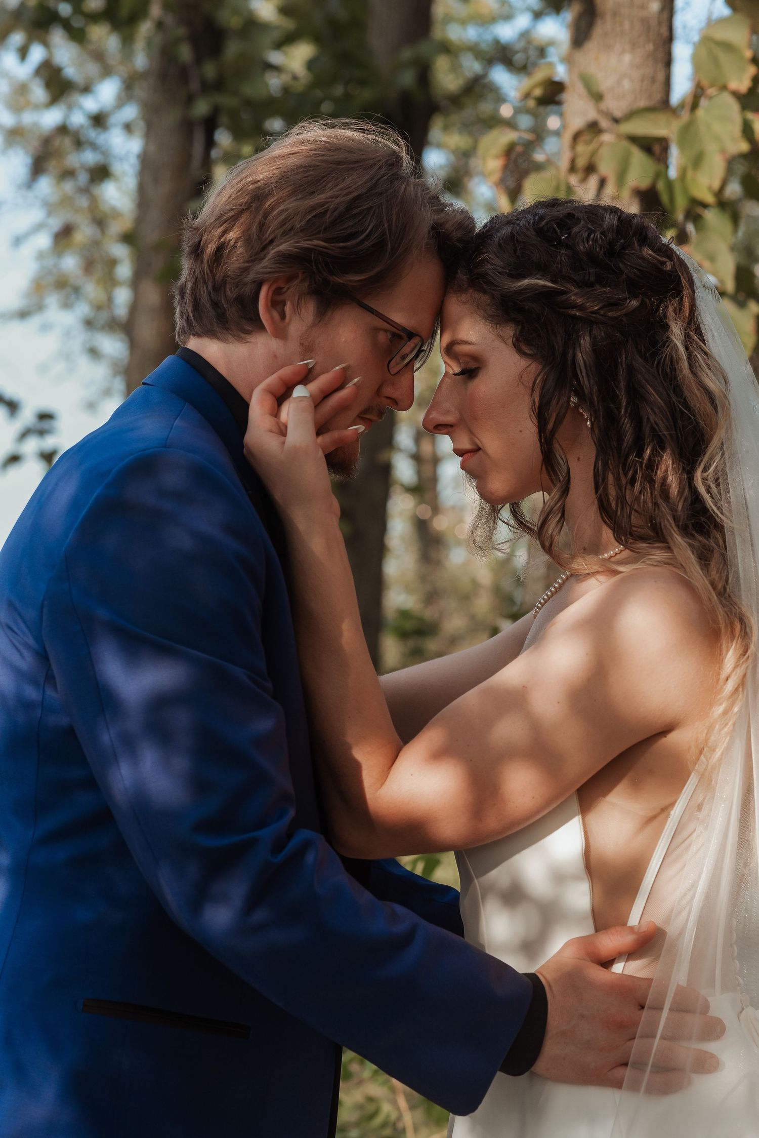 Bride and groom sharing intimate forehead touch moment outdoors in soft dappled sunlight.