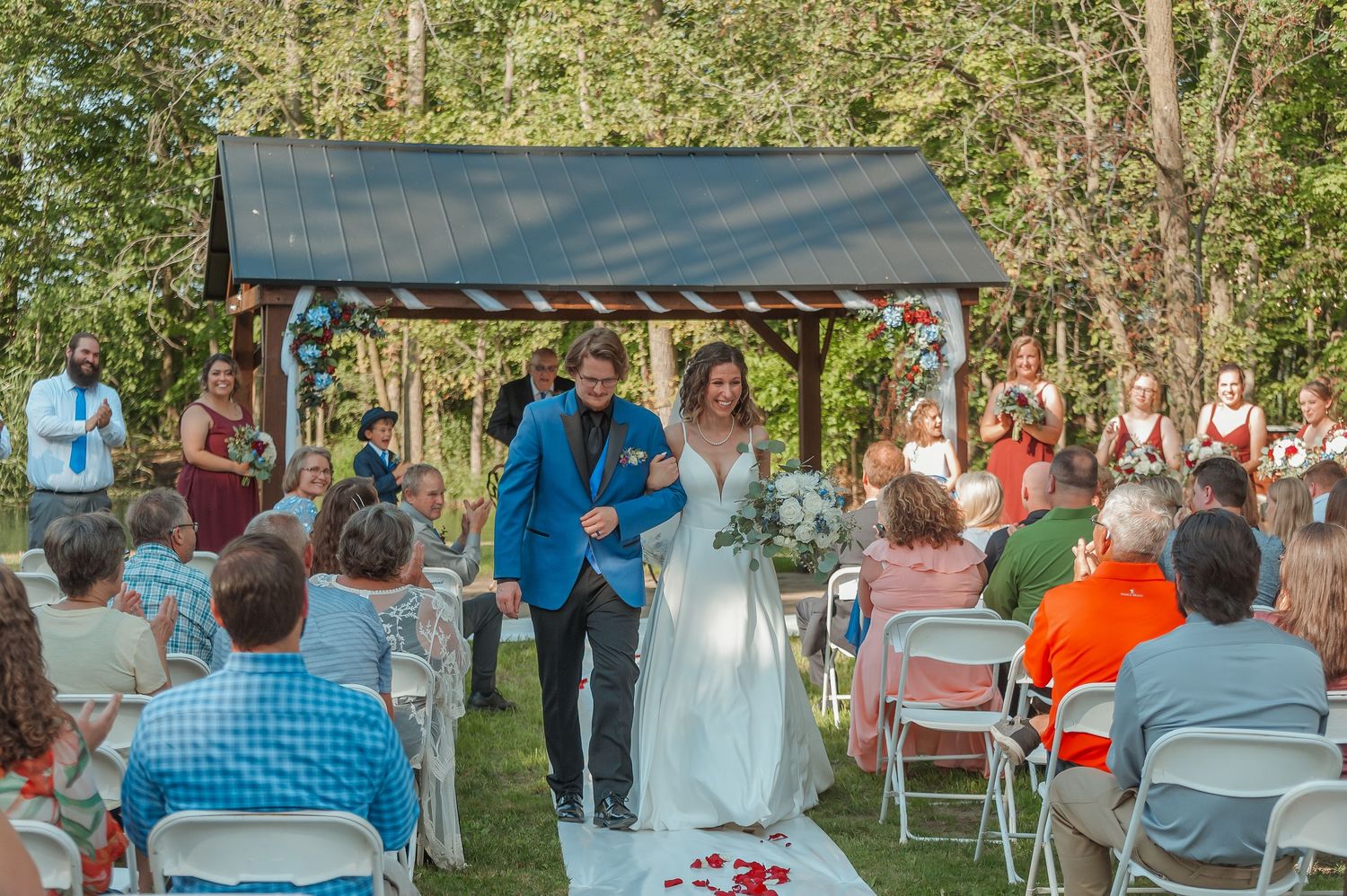 Newlywed couple walks down the aisle as guests cheer at an outdoor wedding ceremony by a wooden gazebo.