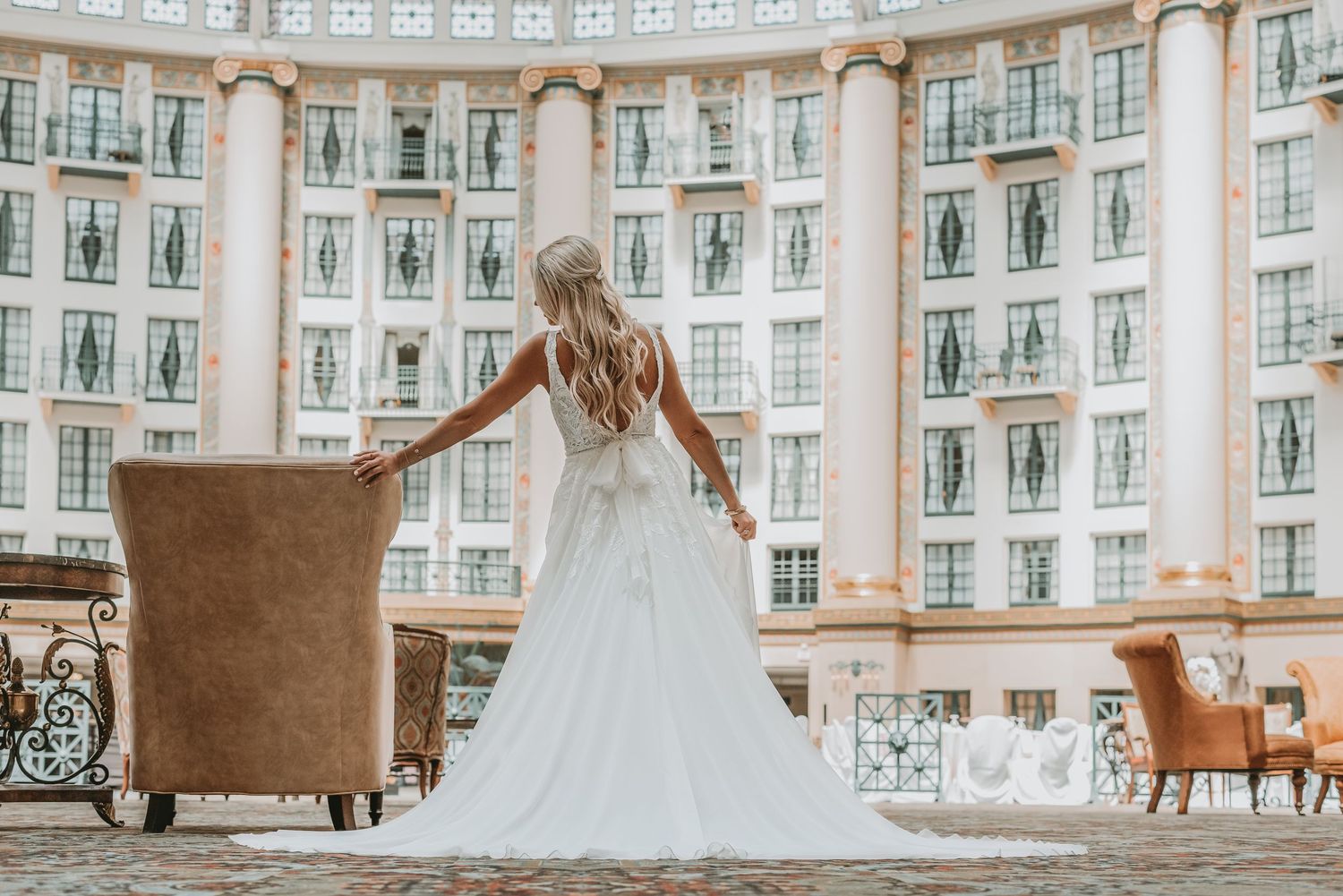 Bride in flowing white wedding gown with long train stands before grand hotel facade with columns.