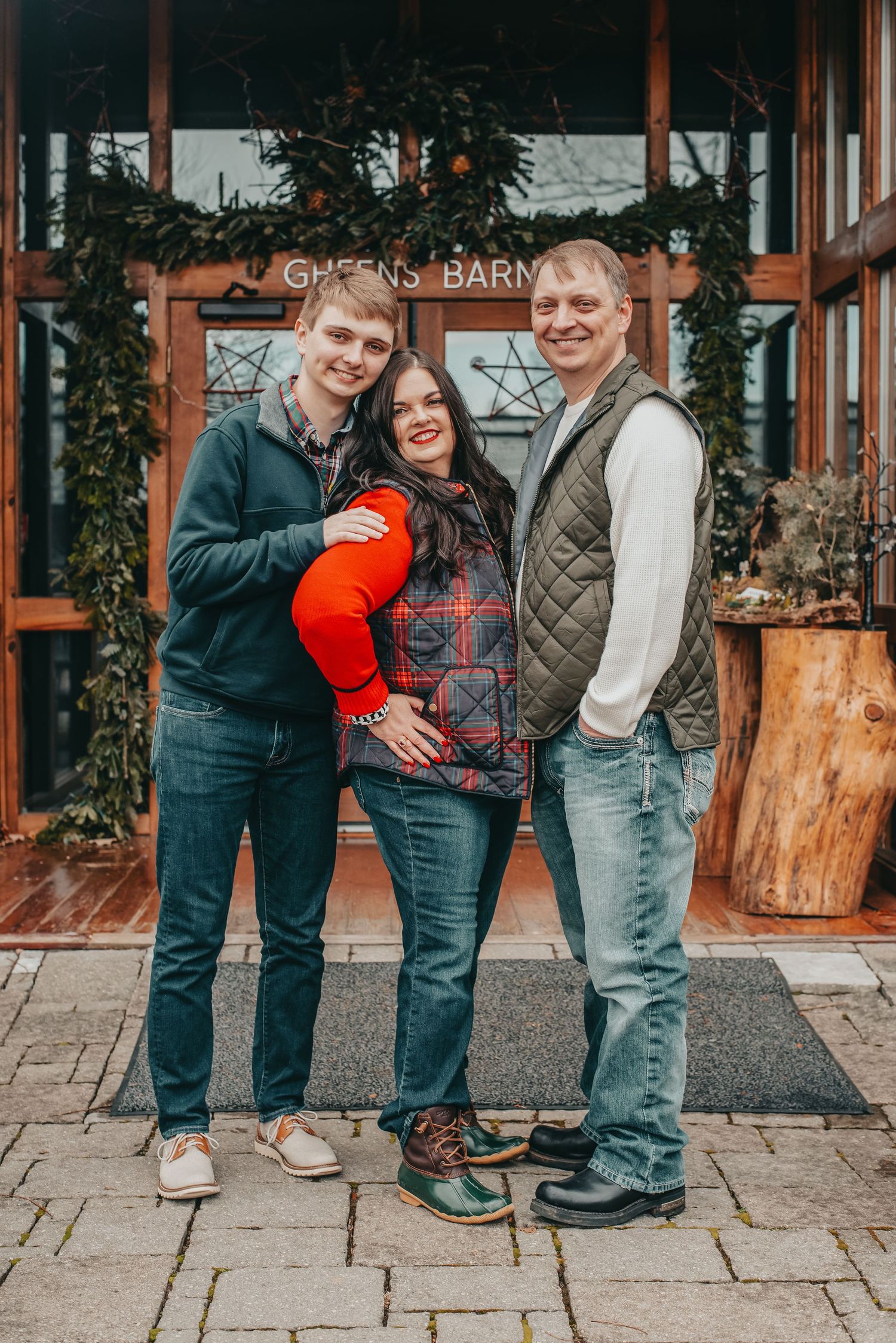 Smiling family of three poses together outside Greens Barn entrance decorated with holiday greenery.
