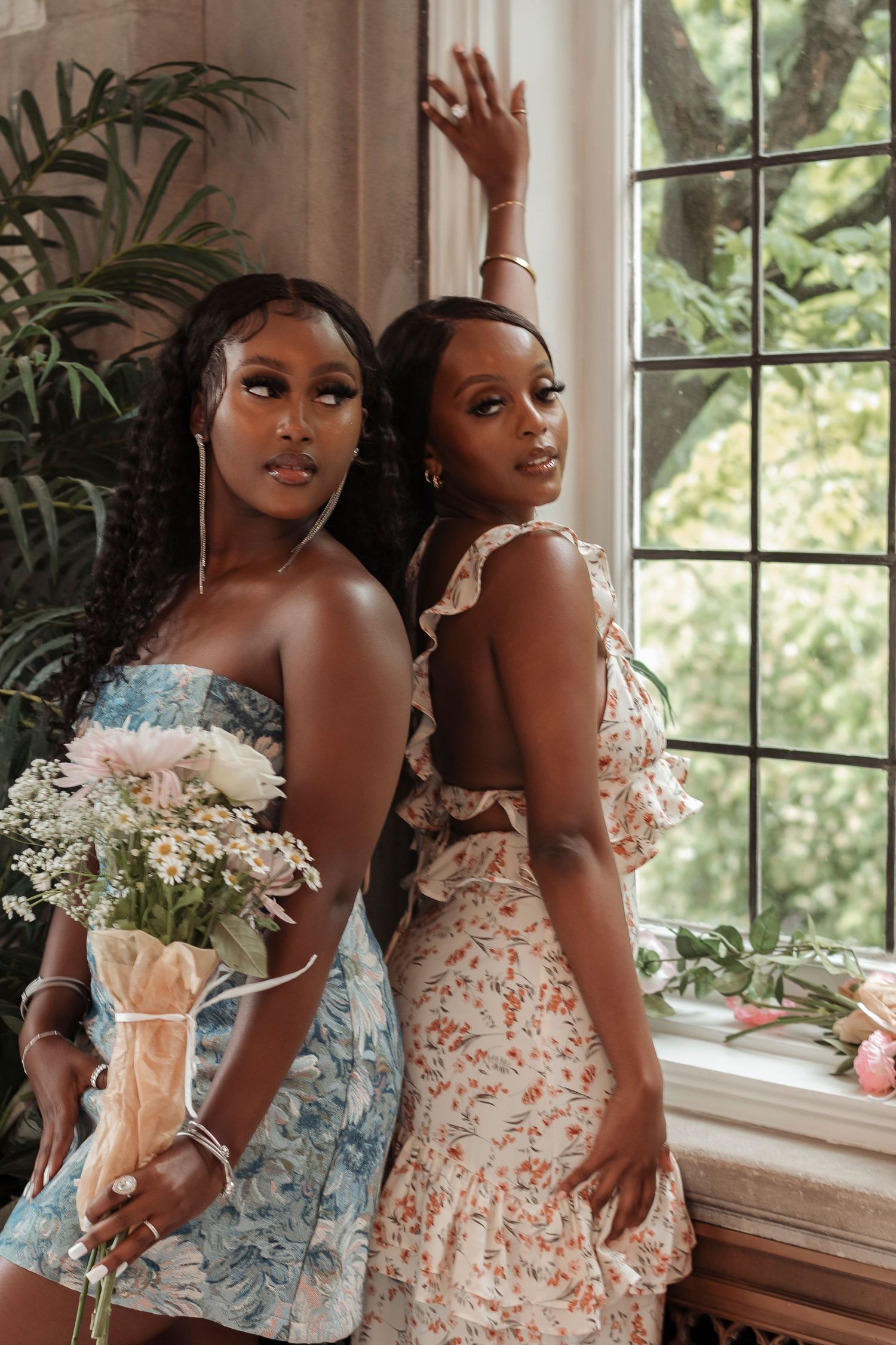 Two young women in floral summer dresses posing by large window with bouquet of flowers.