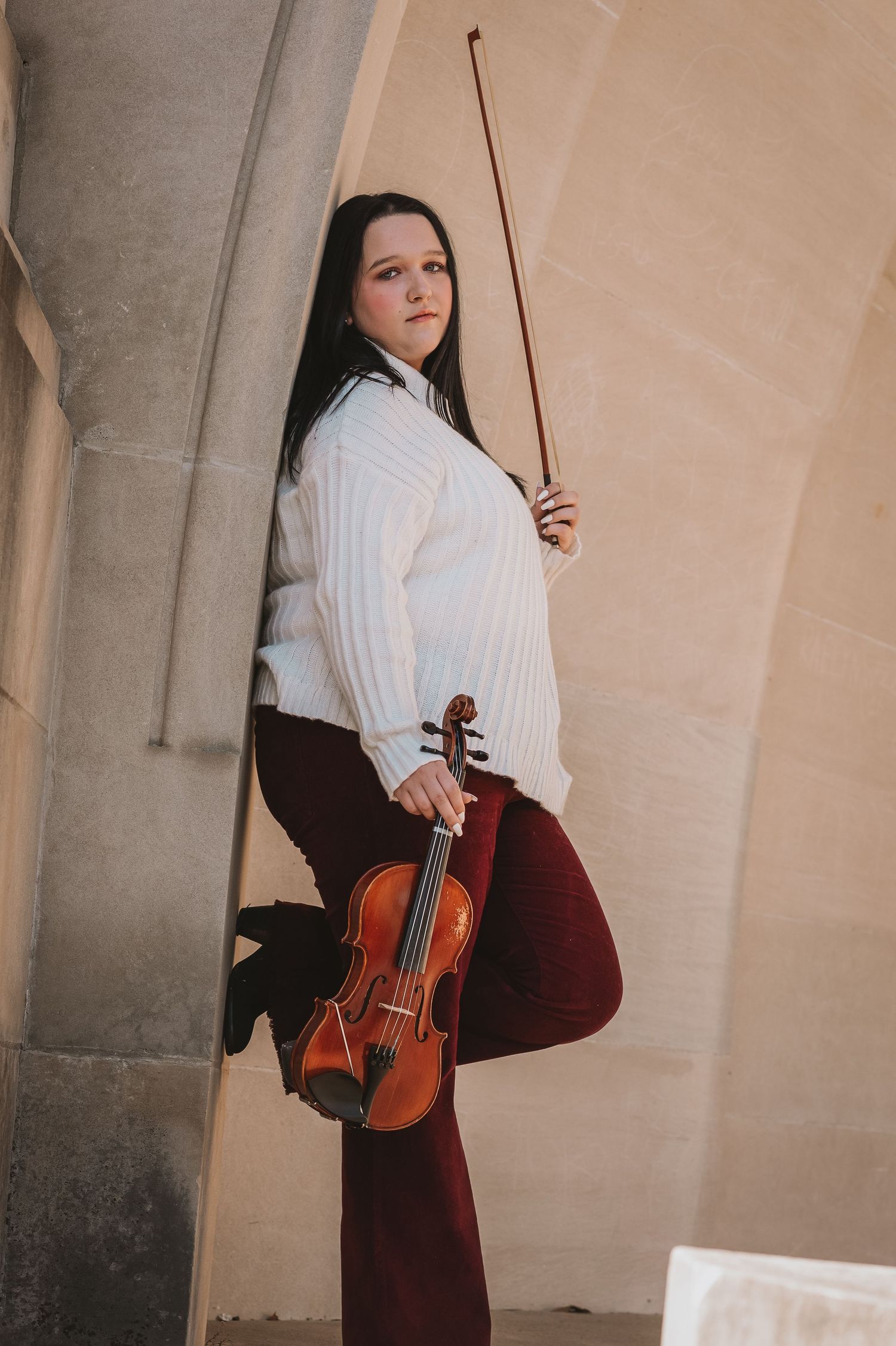 Young woman in white sweater and burgundy pants poses with violin and bow against stone arch wall.