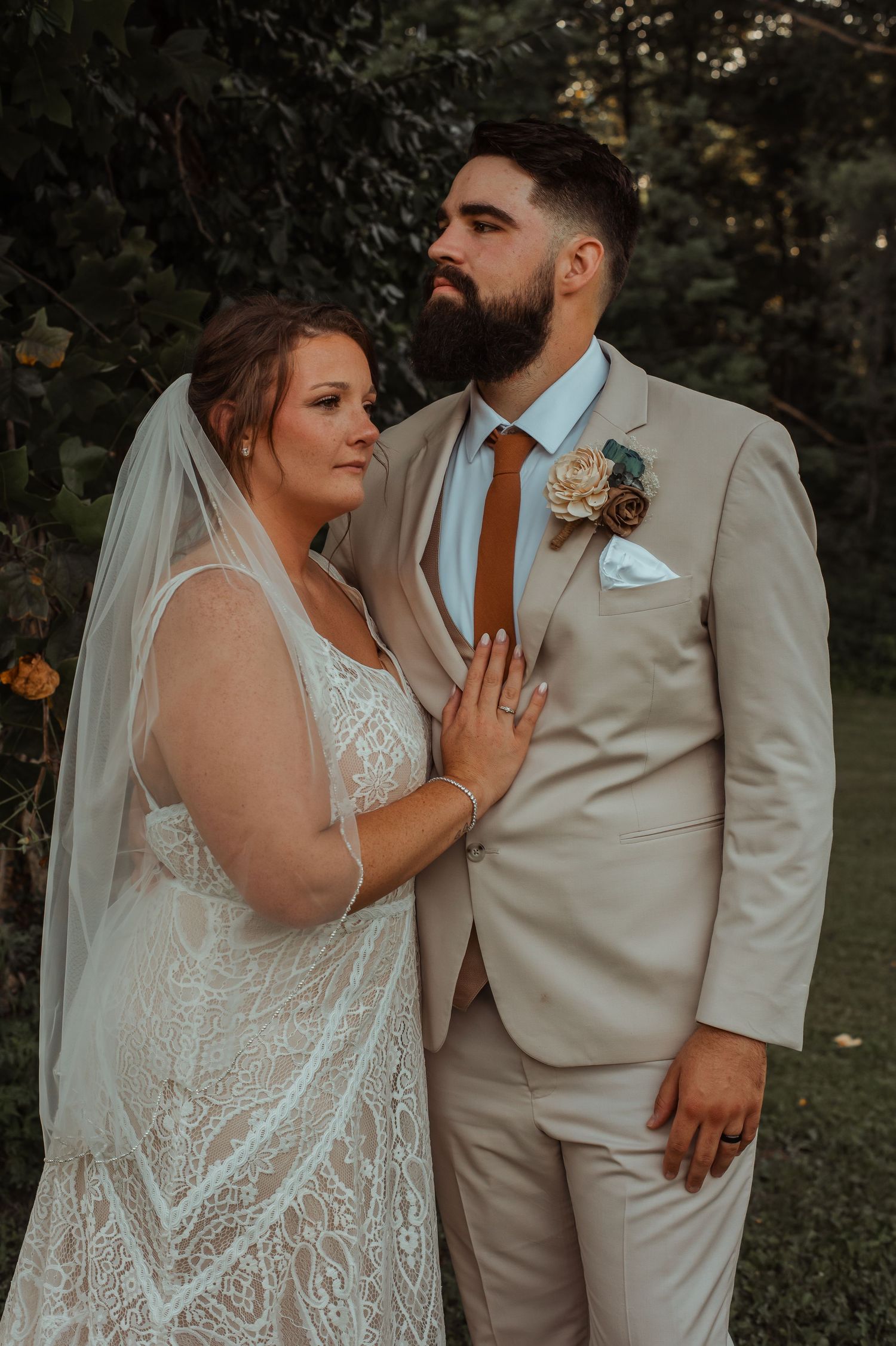 Bride in lace gown with veil and bearded groom in tan suit pose for romantic outdoor portrait.