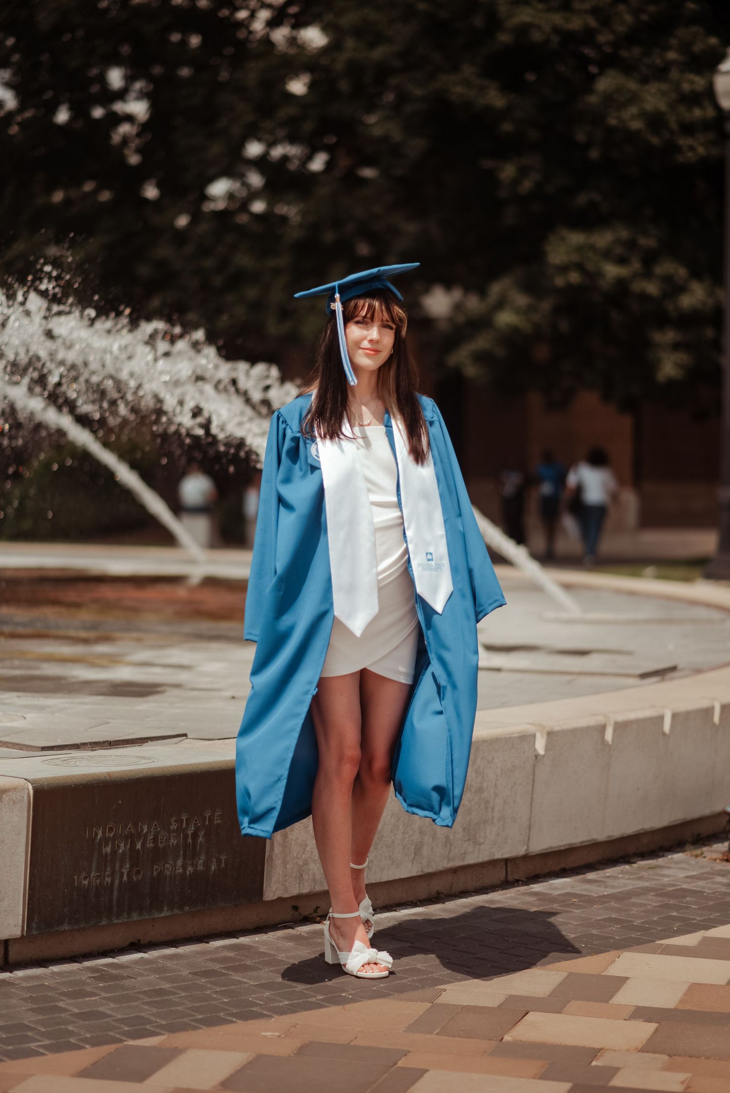 Young woman in blue graduation cap and gown standing near campus fountain wearing white heels and dress.