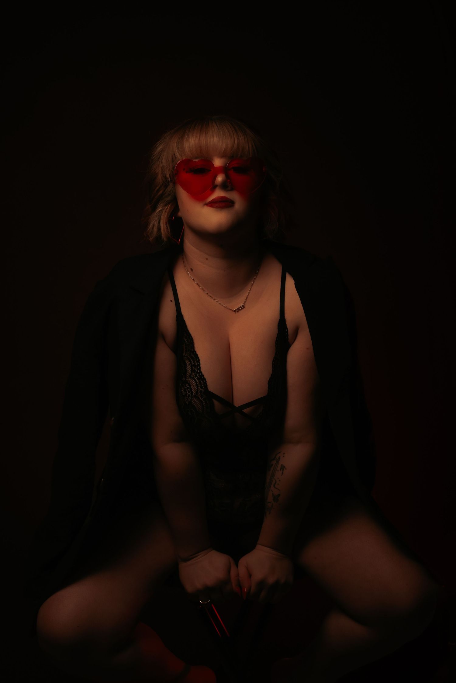 Woman in black lace bodysuit wearing red sunglasses in moody dark lighting.