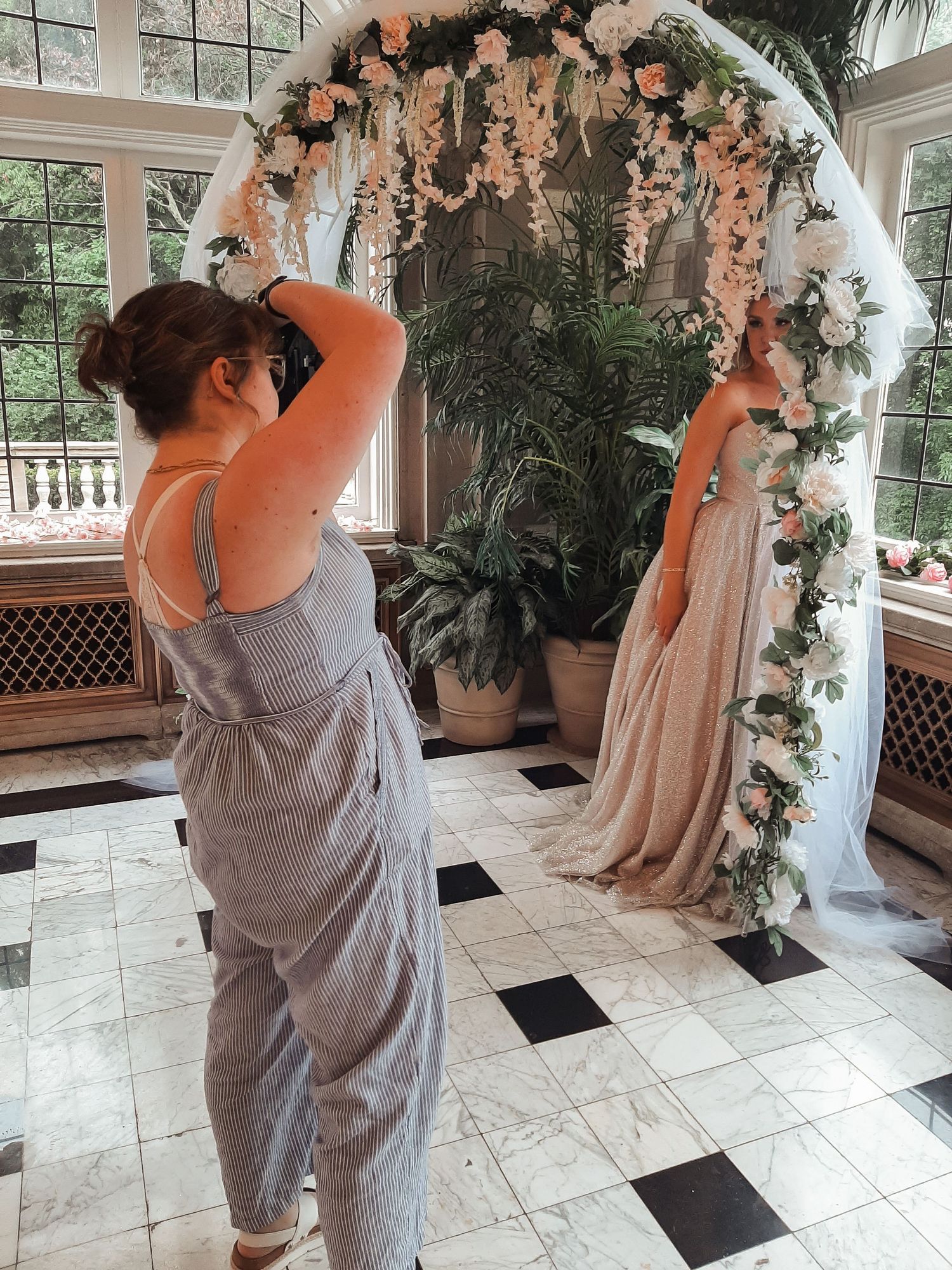 Photographer shooting bride under floral arch in elegant indoor venue.