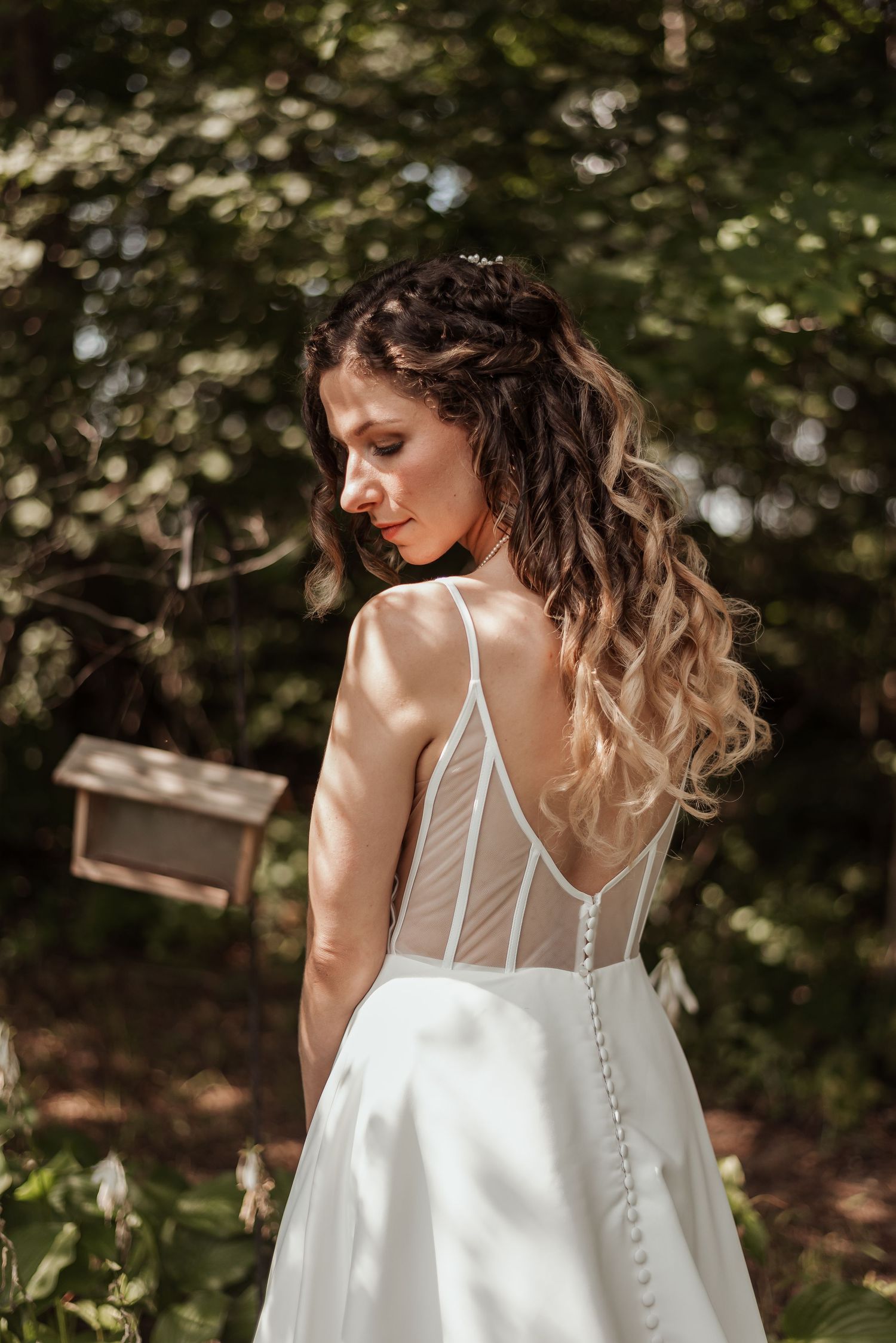 Bride in backless wedding gown with sheer corset detail standing among green trees in sunlight.