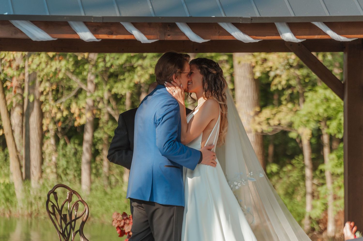 Bride and groom share their first kiss under a rustic wooden gazebo at an outdoor wedding ceremony.