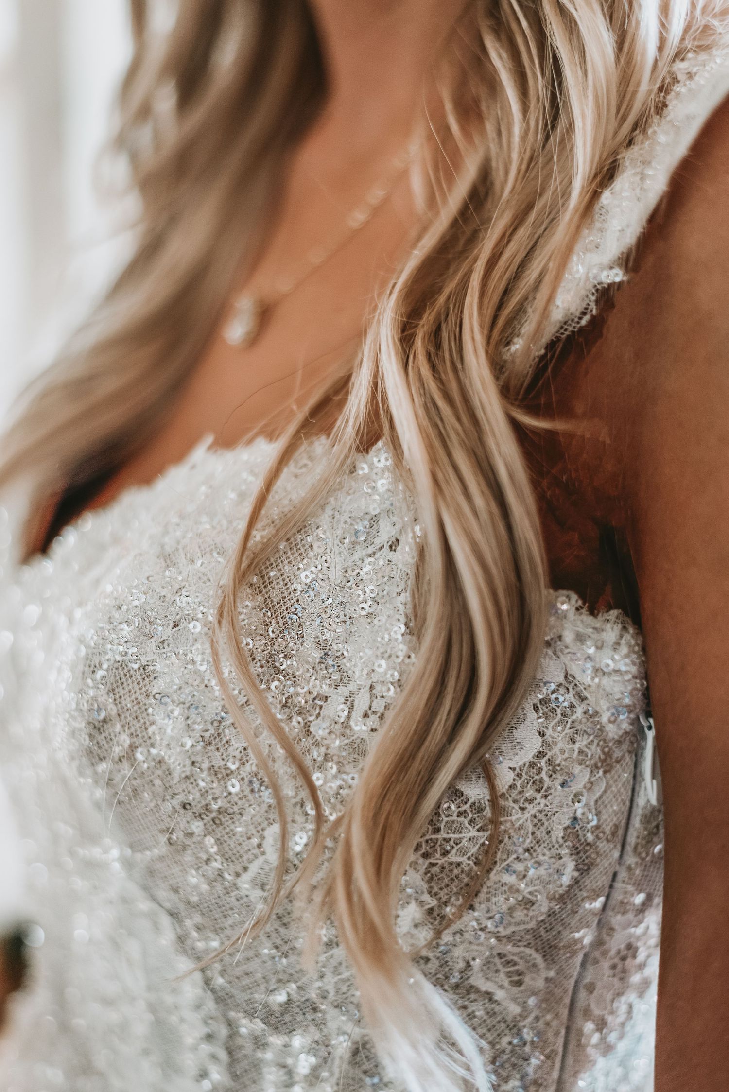 Close-up of a bride's sequined lace wedding dress bodice with flowing blonde hair and delicate necklace.