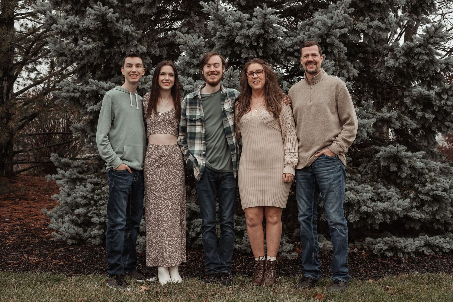 Five smiling family members pose outdoors in front of evergreen trees during a fall family portrait session.