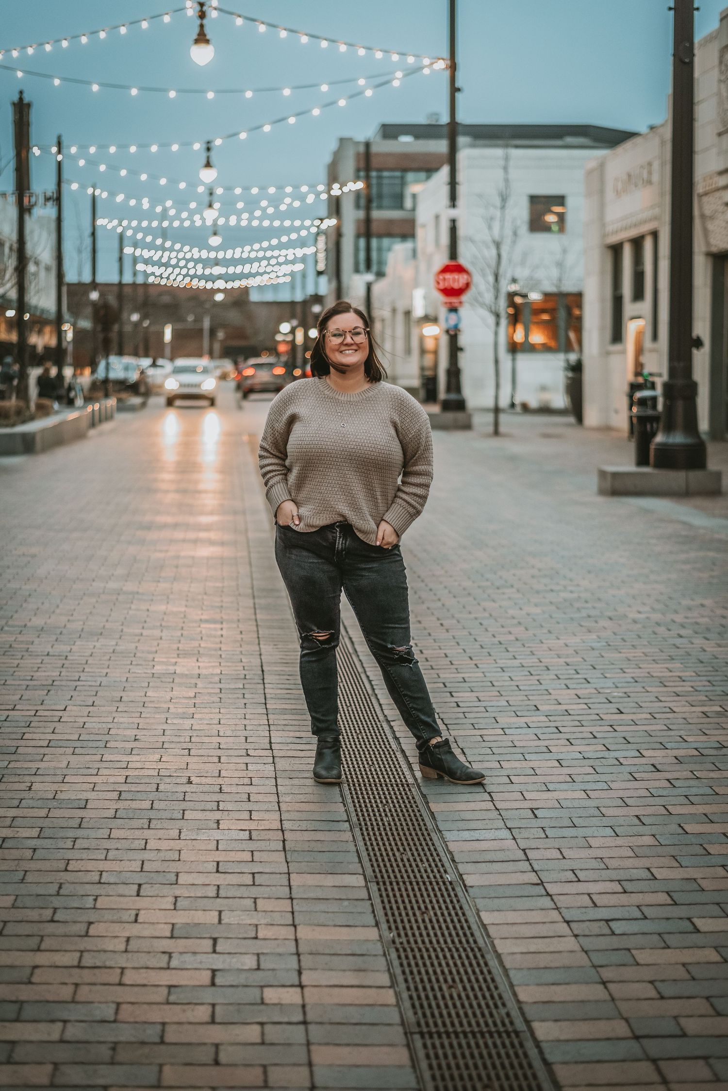 Smiling woman in beige sweater and ripped jeans standing on brick downtown street under string lights.