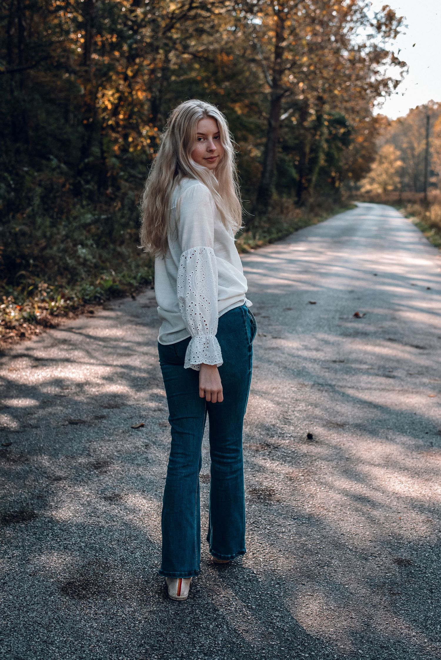 Blonde woman in white blouse and flared jeans standing on rural road lined with autumn trees.