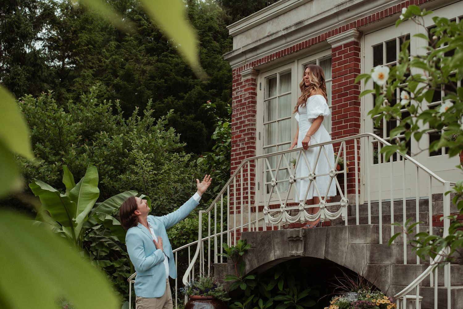 Romantic couple recreating Romeo and Juliet scene at a brick mansion, man reaching up to woman on balcony.